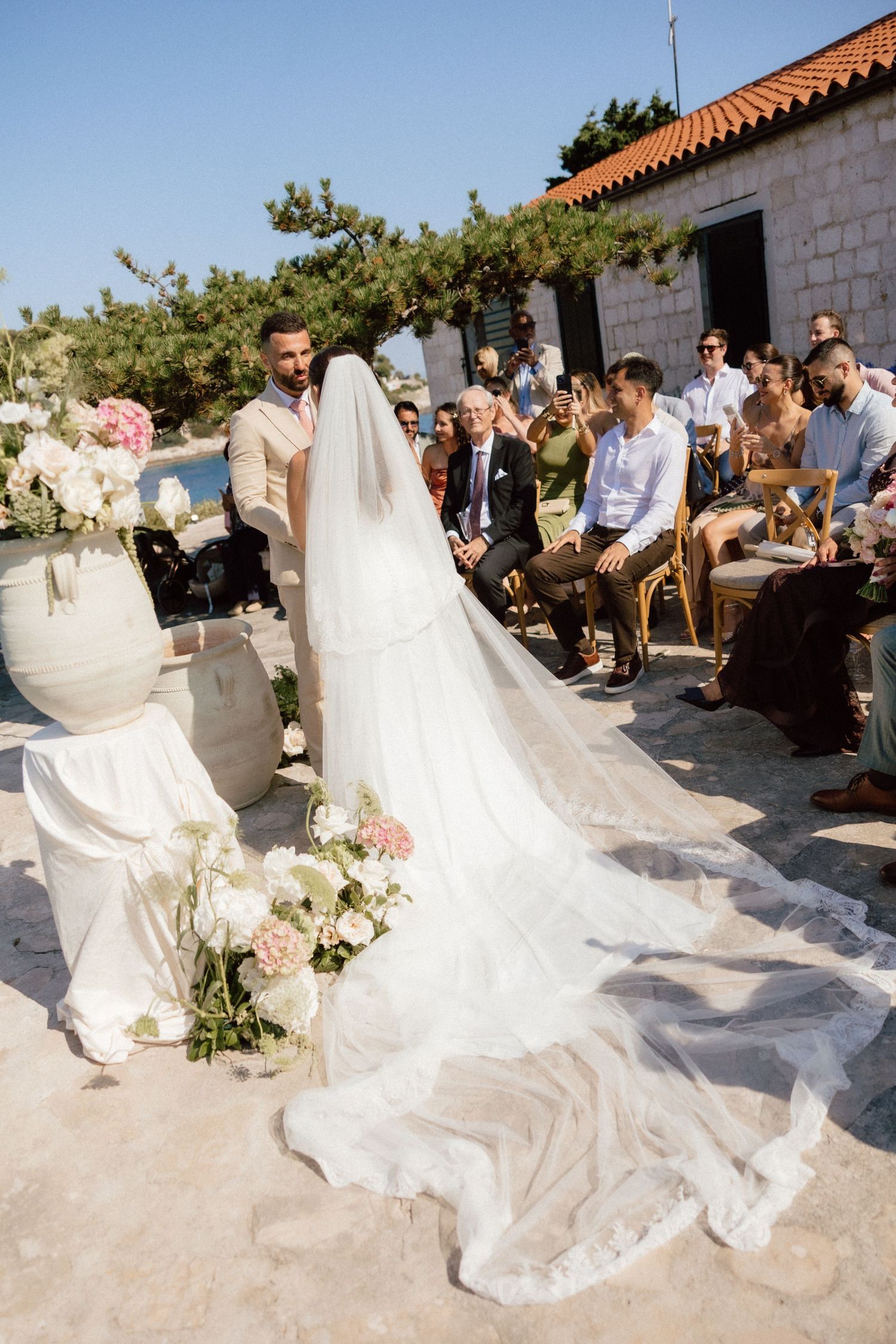 Bride in flowing veil walks toward groom during outdoor seaside wedding ceremony with guests seated.