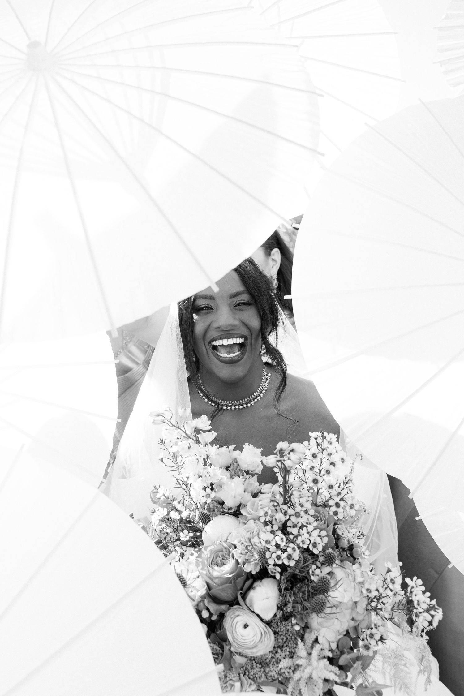 Black and white photo of a joyful bride laughing, holding a floral bouquet, surrounded by white parasols on her wedding day.