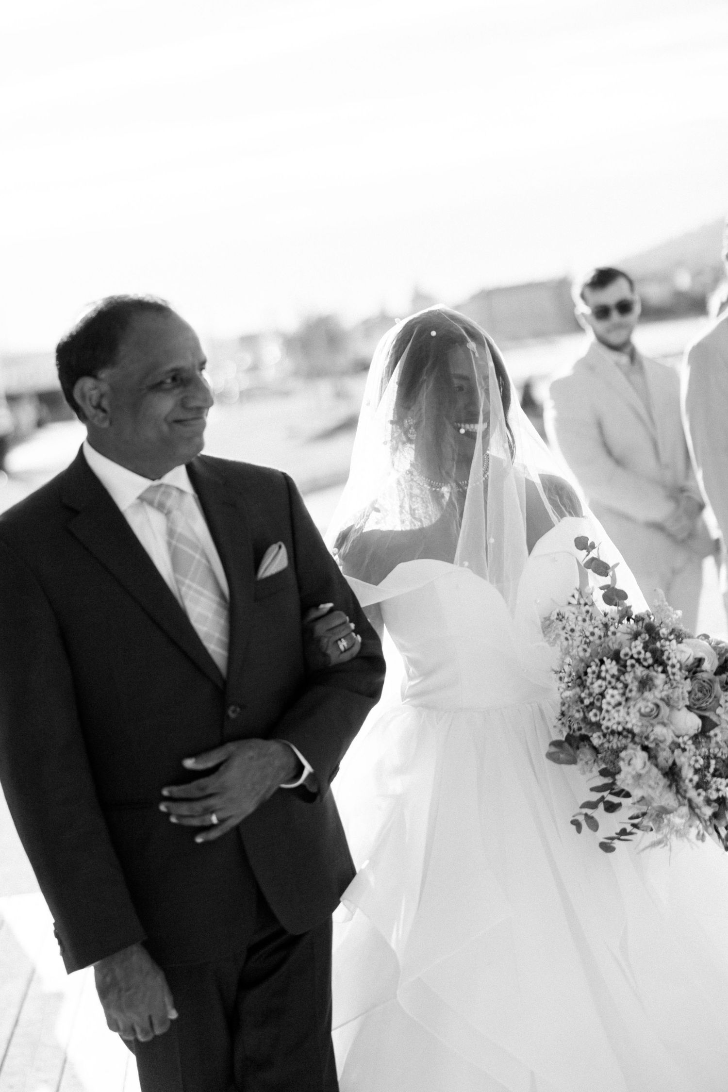 Black and white photo of a bride in a veil holding flowers walking with an older man at a wedding.