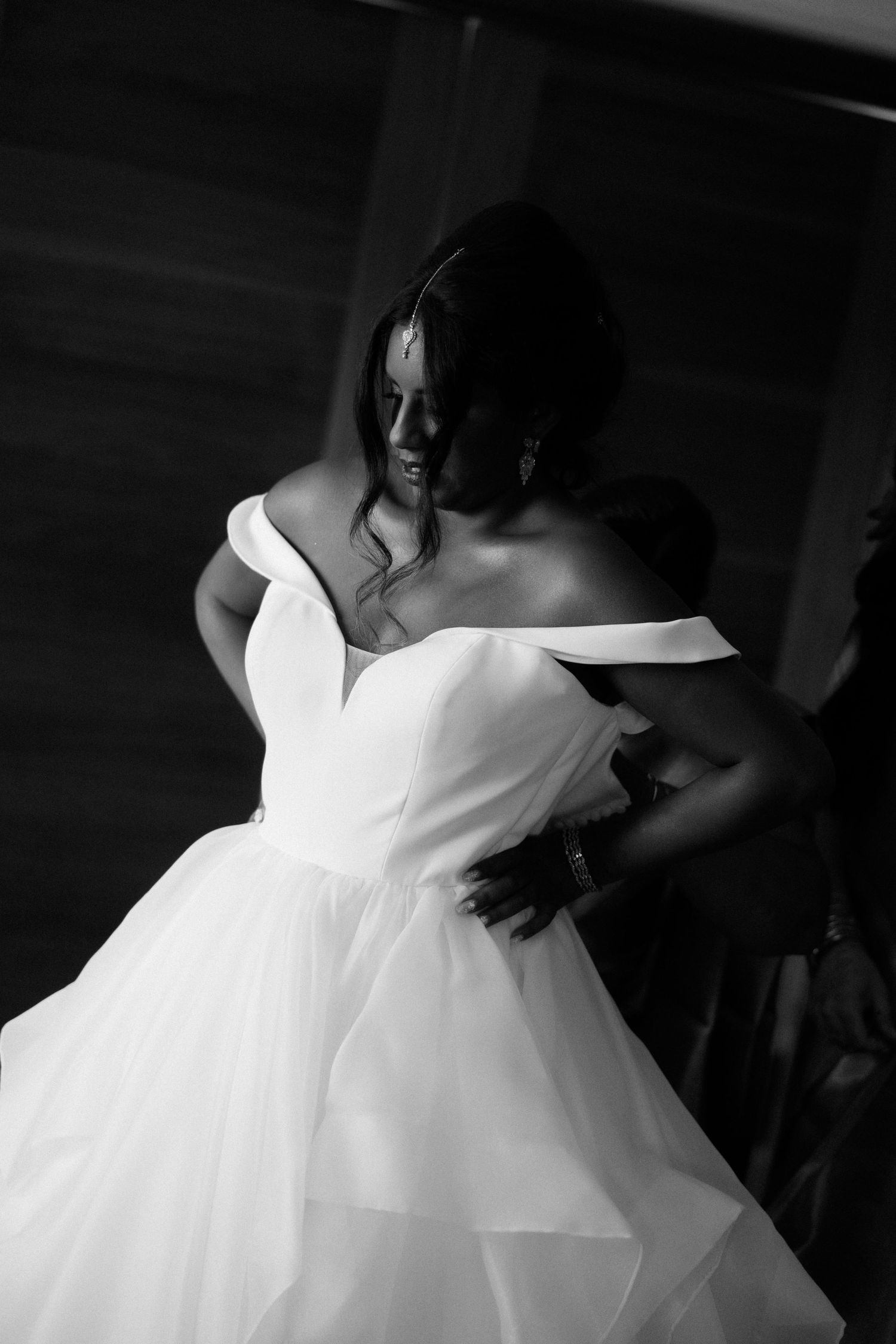 Black and white photo of a bride in an off-shoulder ballgown being helped into her dress by a bridesmaid.