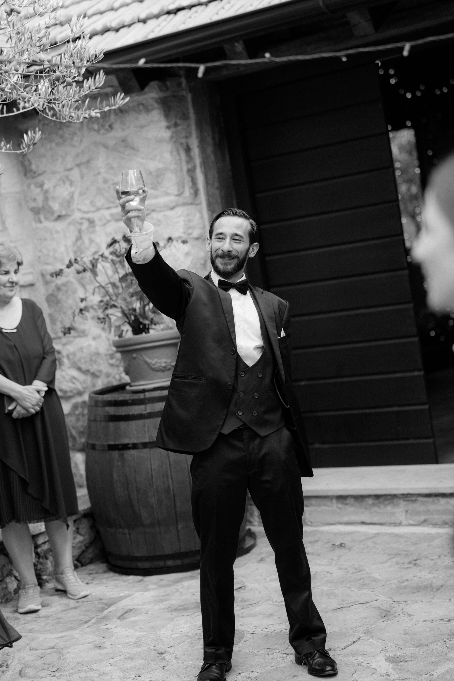 A wedding guest in a tuxedo raises his glass in celebration at an outdoor reception.