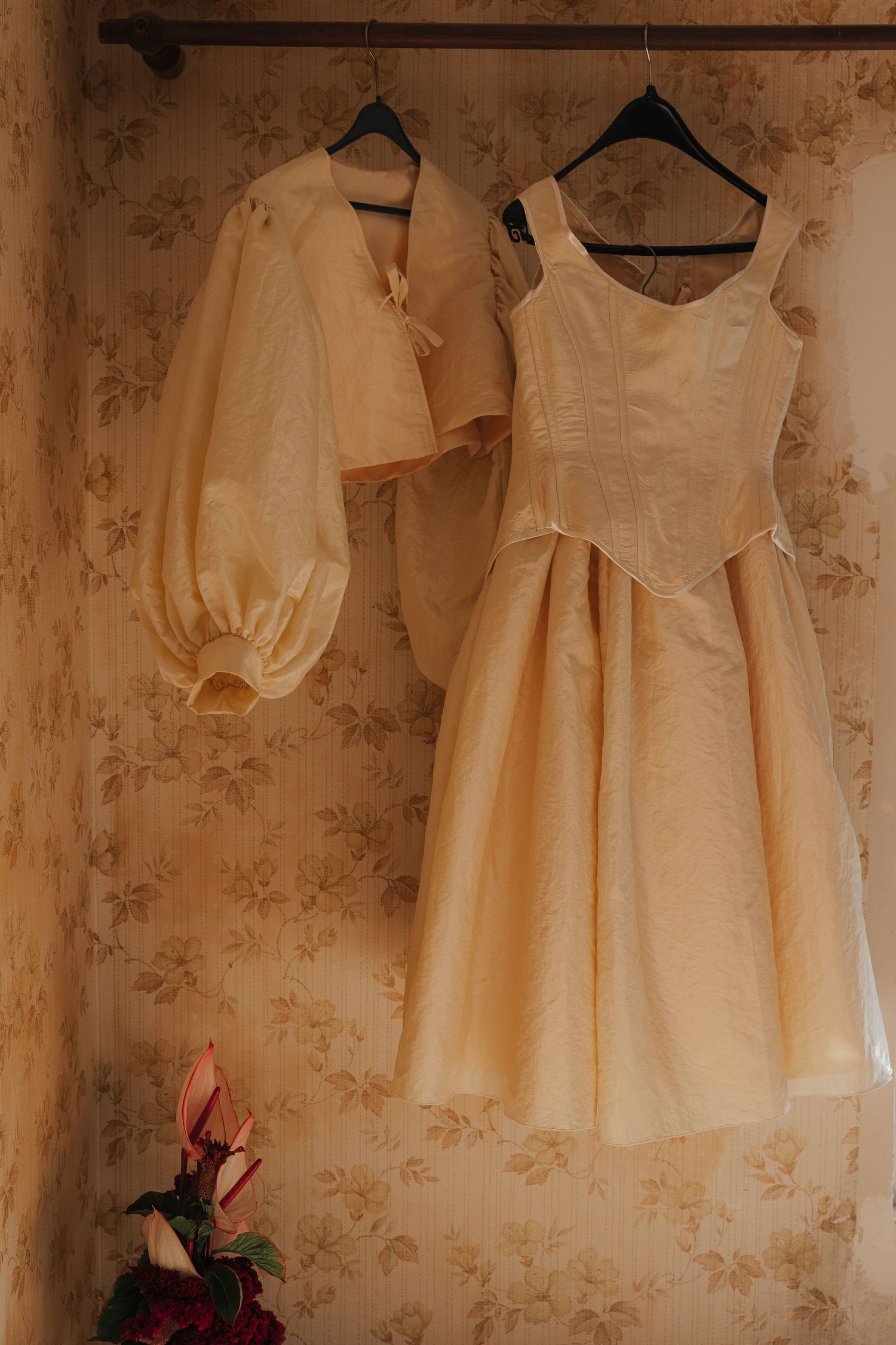 Vintage cream-colored dresses hanging on black hangers against floral wallpaper in a dimly lit room.