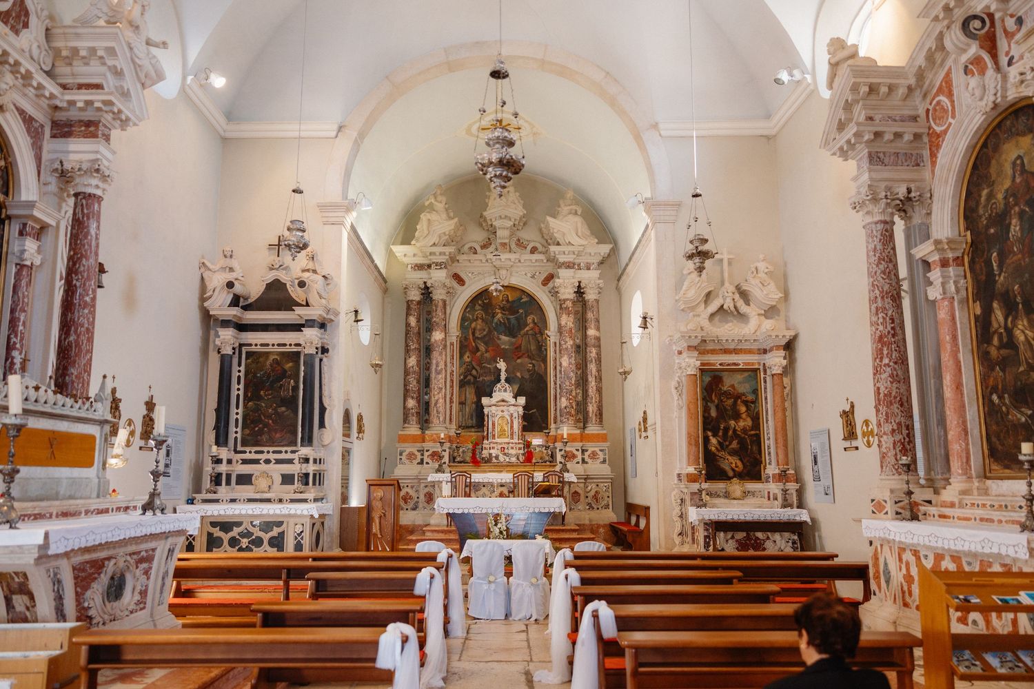 Interior panorama of an ornate Baroque church with white walls, gilded altars, and wooden pews in a row.