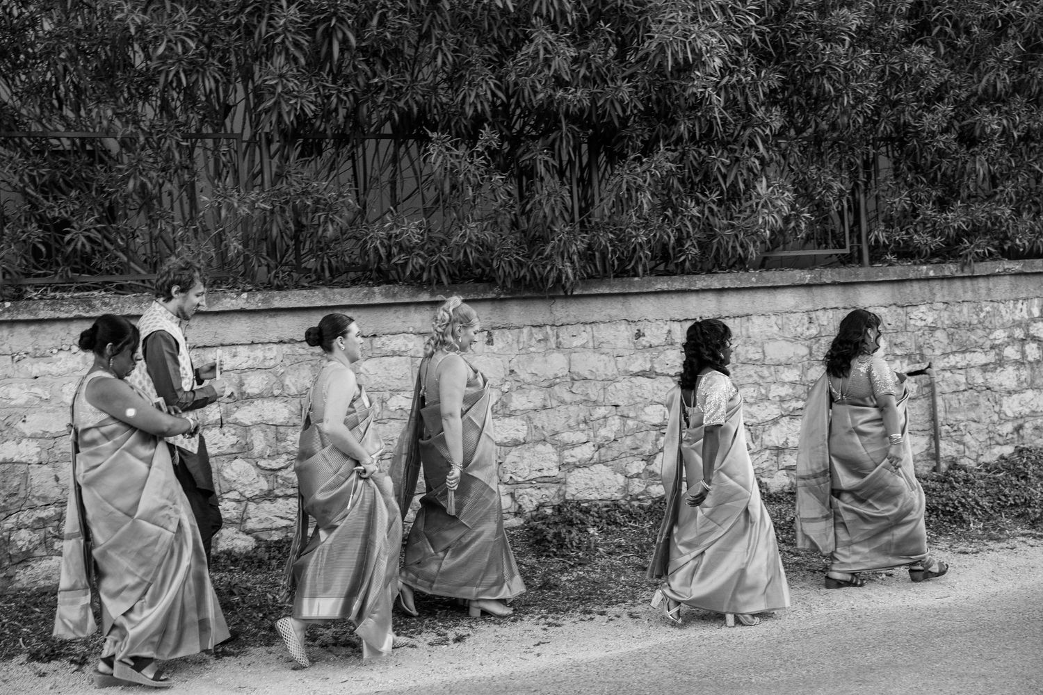 Black and white photo of bridesmaids in sarees walking along stone wall path.