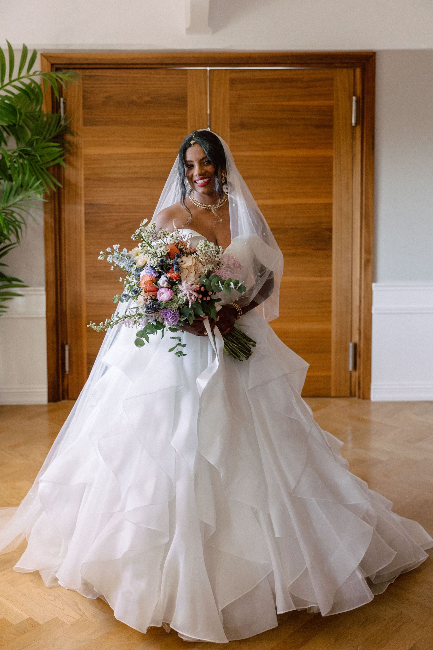 Radiant bride in off-shoulder layered white ball gown holding colorful bridal bouquet with veil.