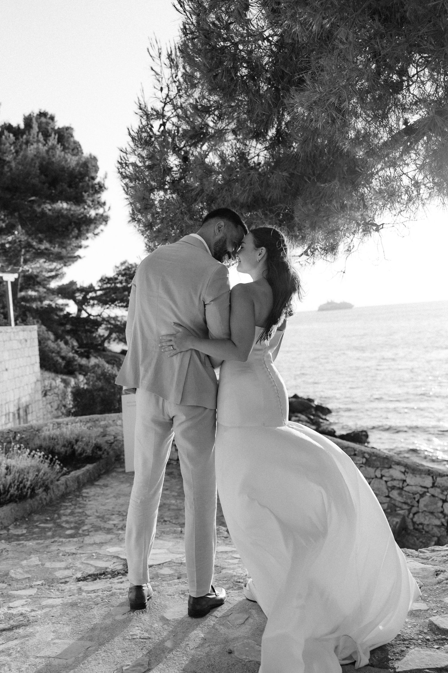 Black and white photo of bride and groom kissing by the sea under a pine tree.