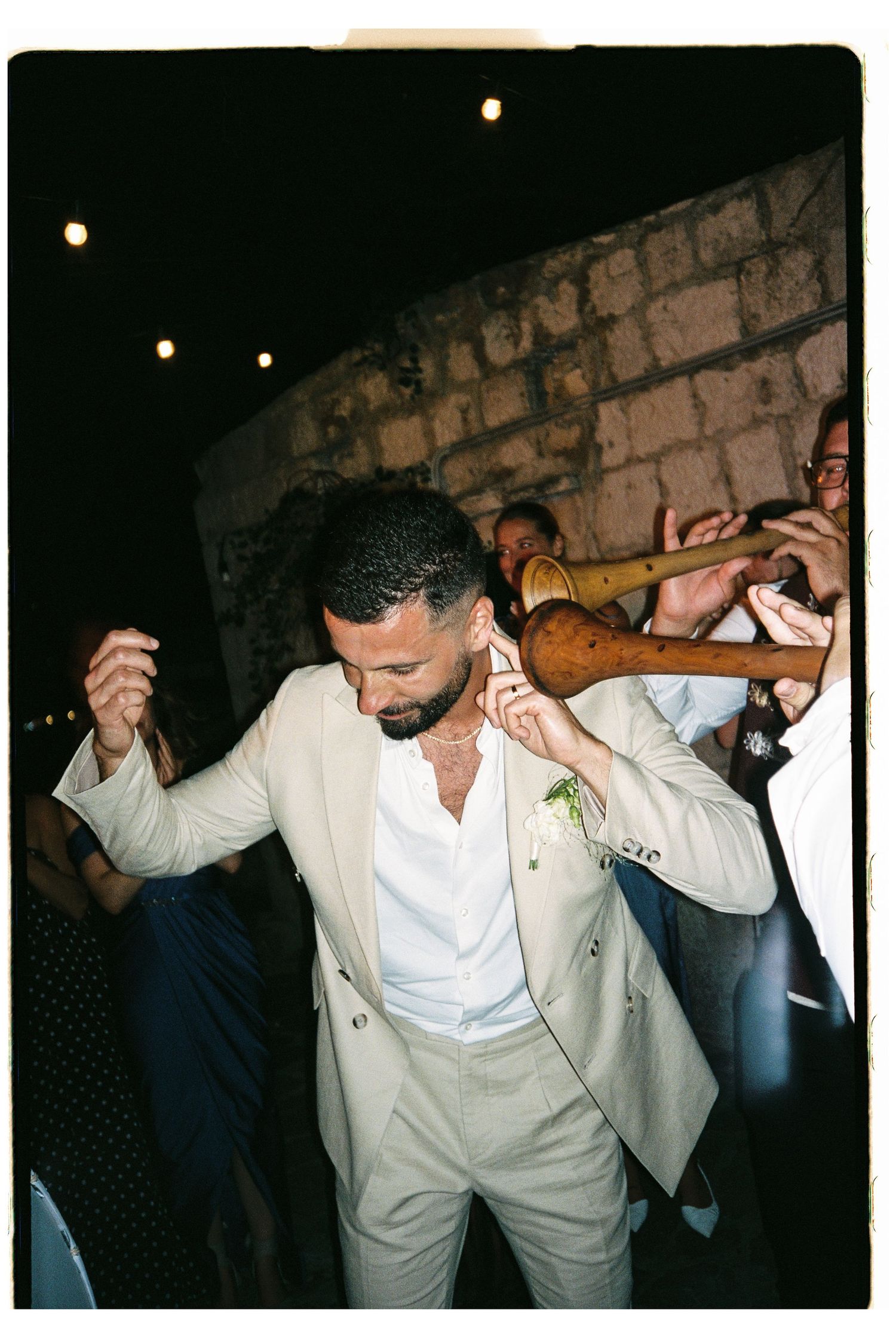 Groom in cream suit dancing energetically at wedding reception with stone walls and string lights.