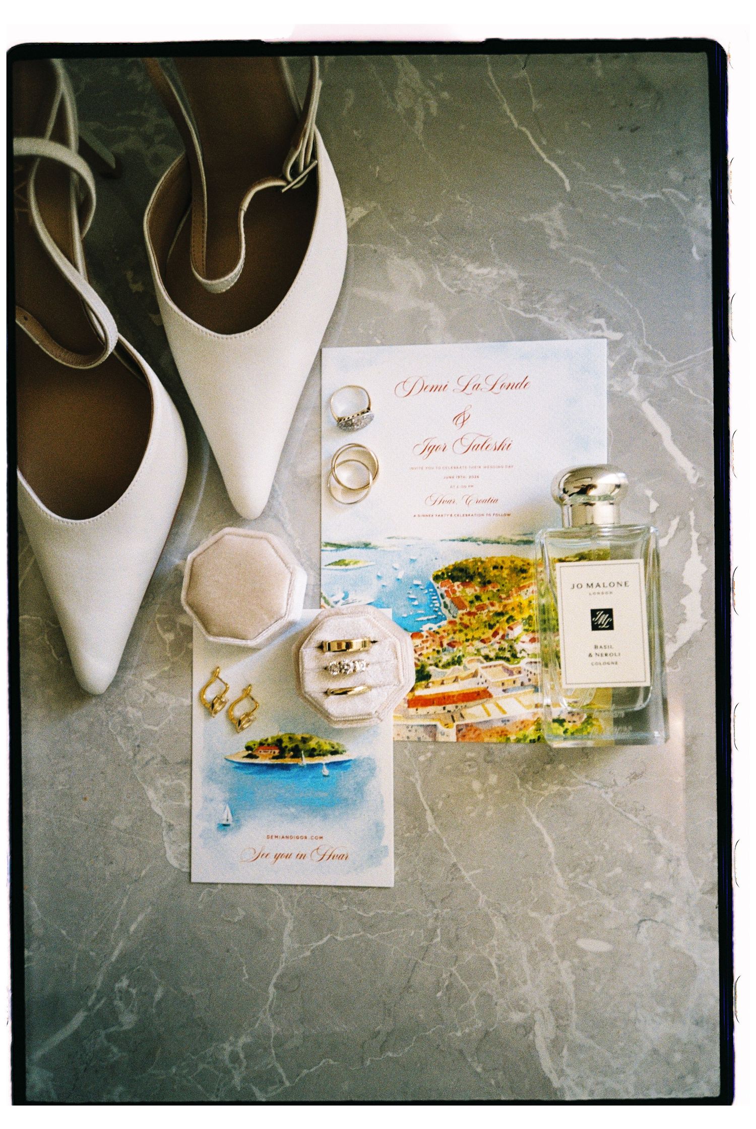 Elegant wedding flat lay with white heels, Jo Malone perfume, gold rings, invitation, and floral stationery on marble.