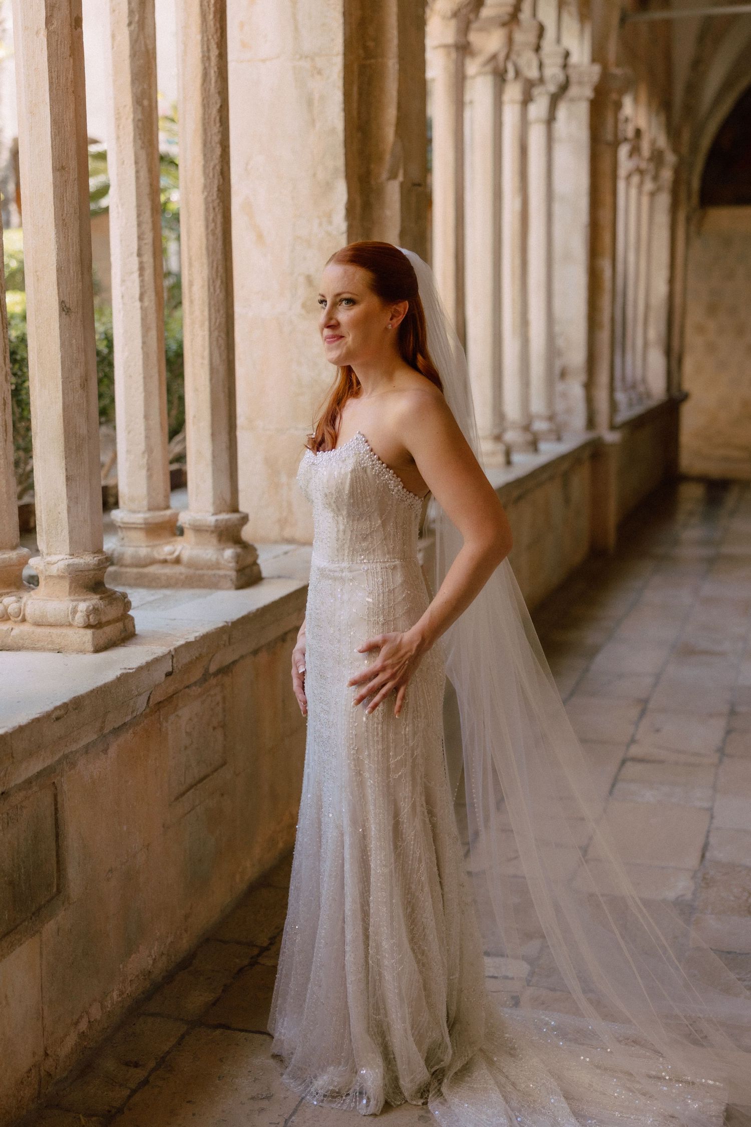 Bride posing in a lacey white gown along a beautiful stone colonnade with natural lighting  in Dubrovnik, Croatia.