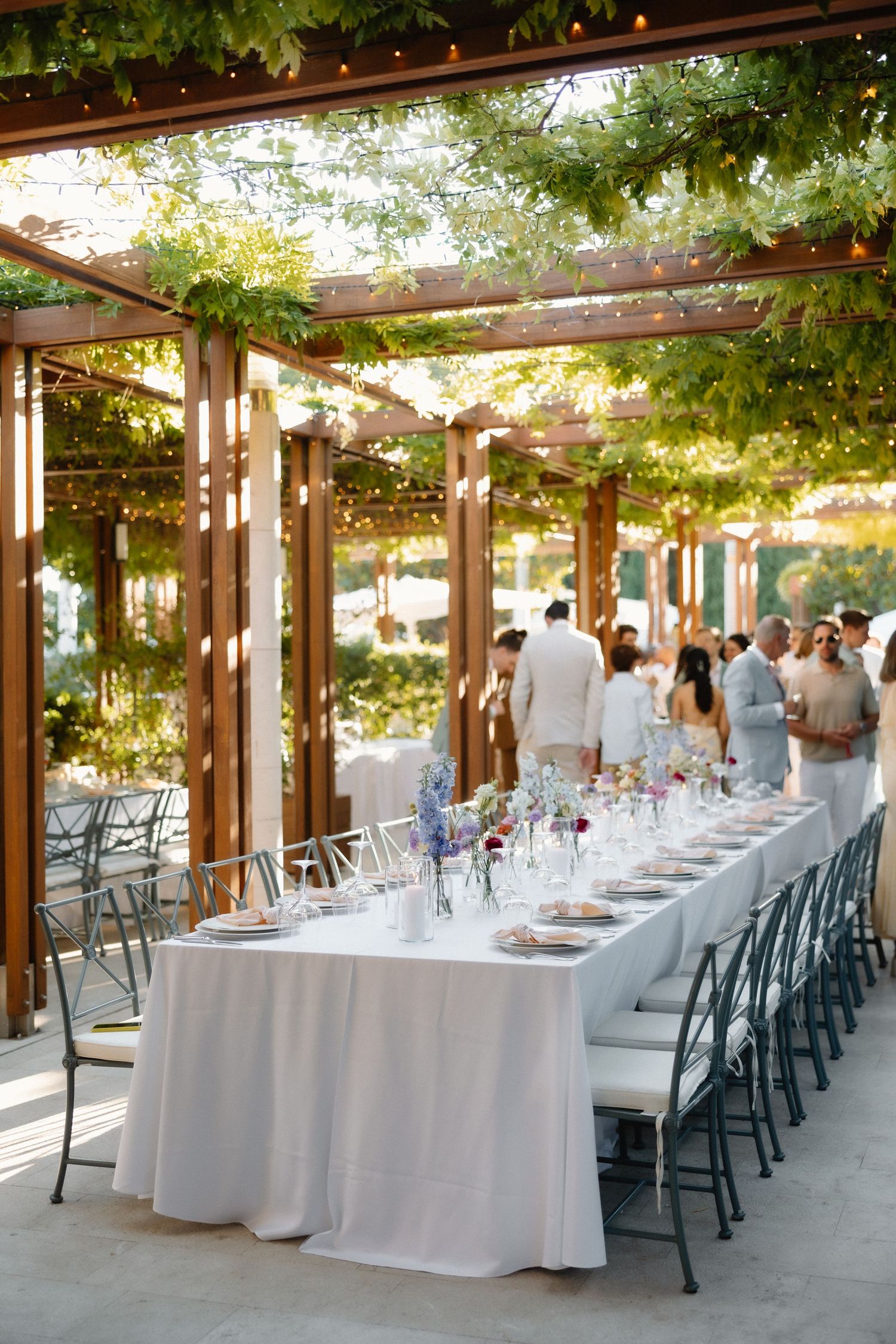 Elegant outdoor wedding reception with long white-draped table, floral centerpieces, string lights, and wooden pergola covered in greenery.