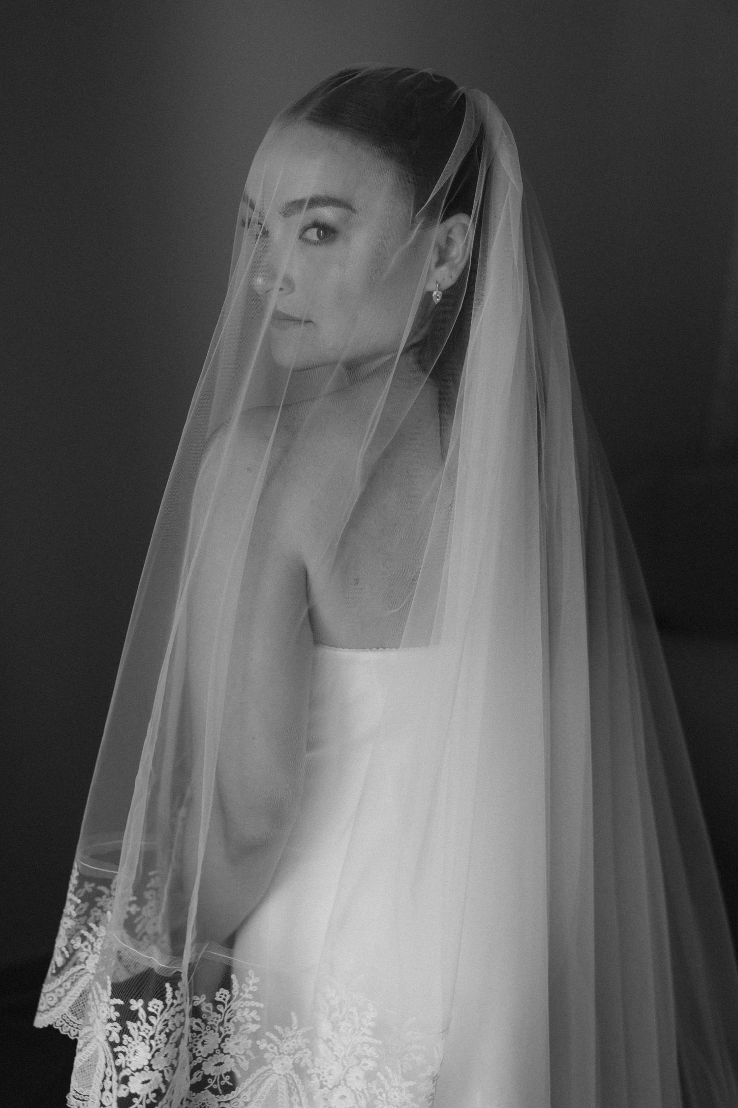 Black and white portrait of a bride wearing a lace wedding dress and flowing veil, gazing elegantly to the side.