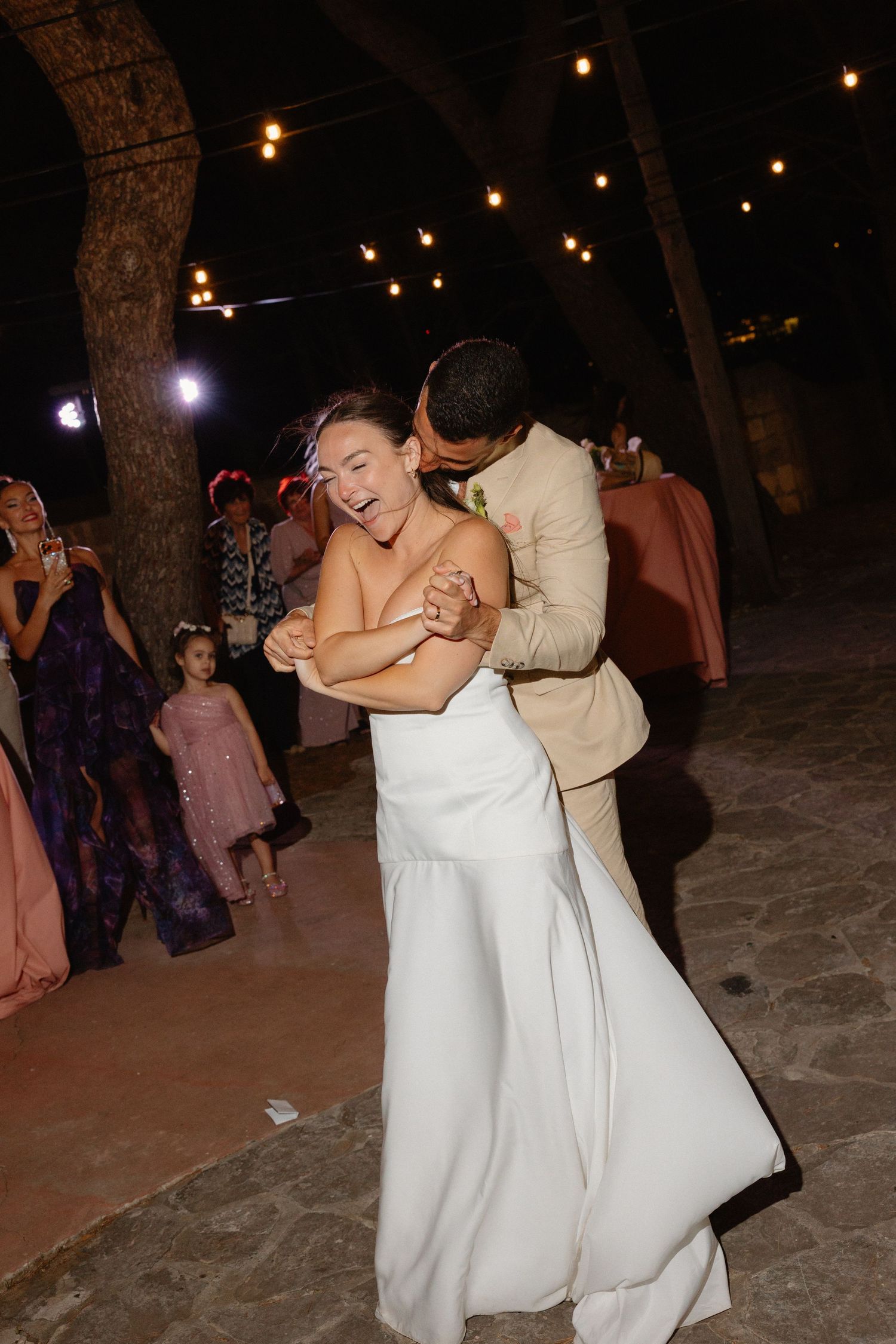 Bride laughing joyfully as groom dips her during first dance under string lights at night.