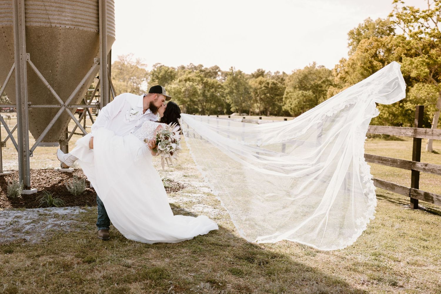 Dramatic wedding photo with bride's veil flowing in the wind near a rustic grain silo.