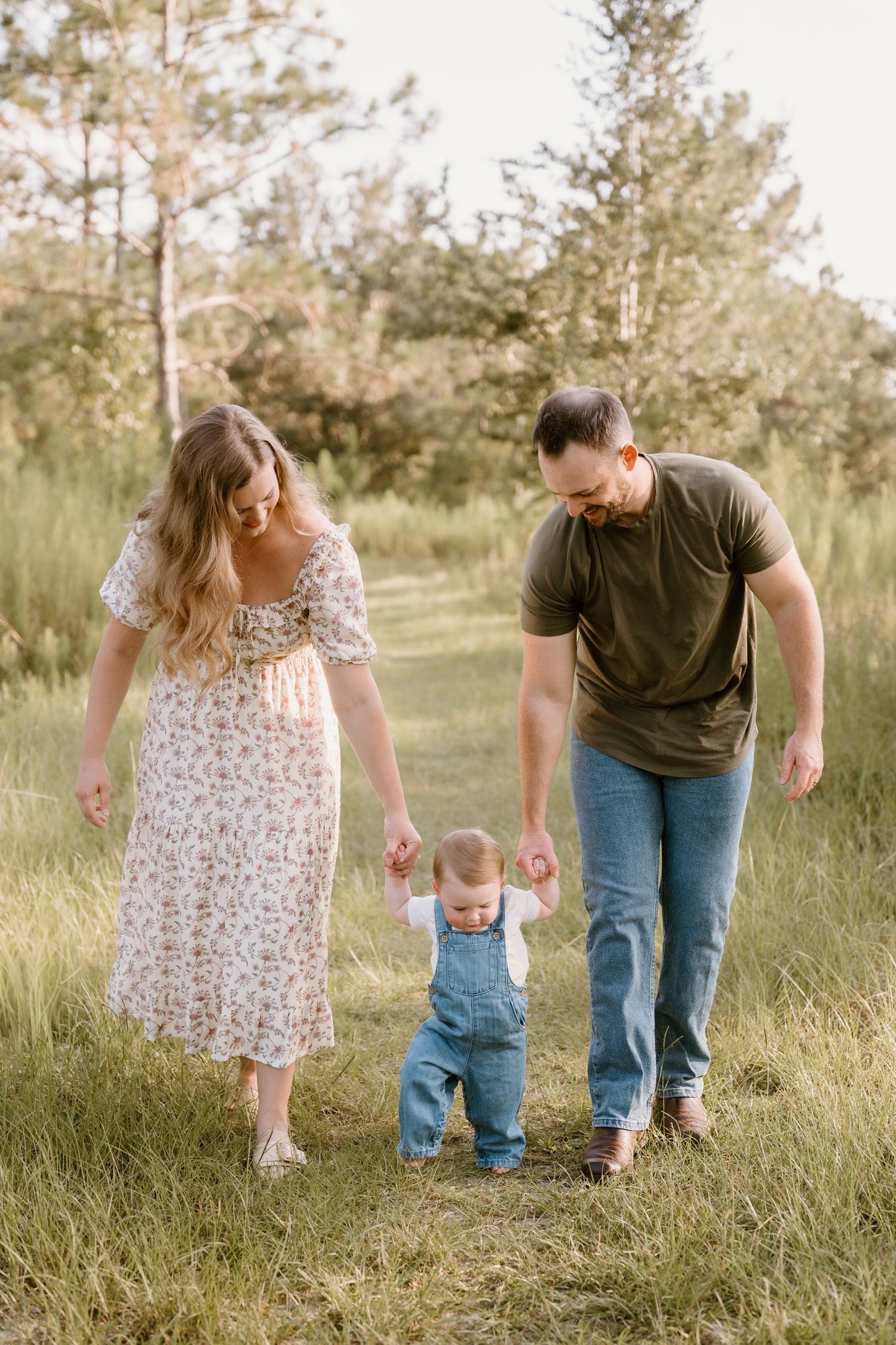 A family walks together through a grassy field, helping their toddler take steps between them.