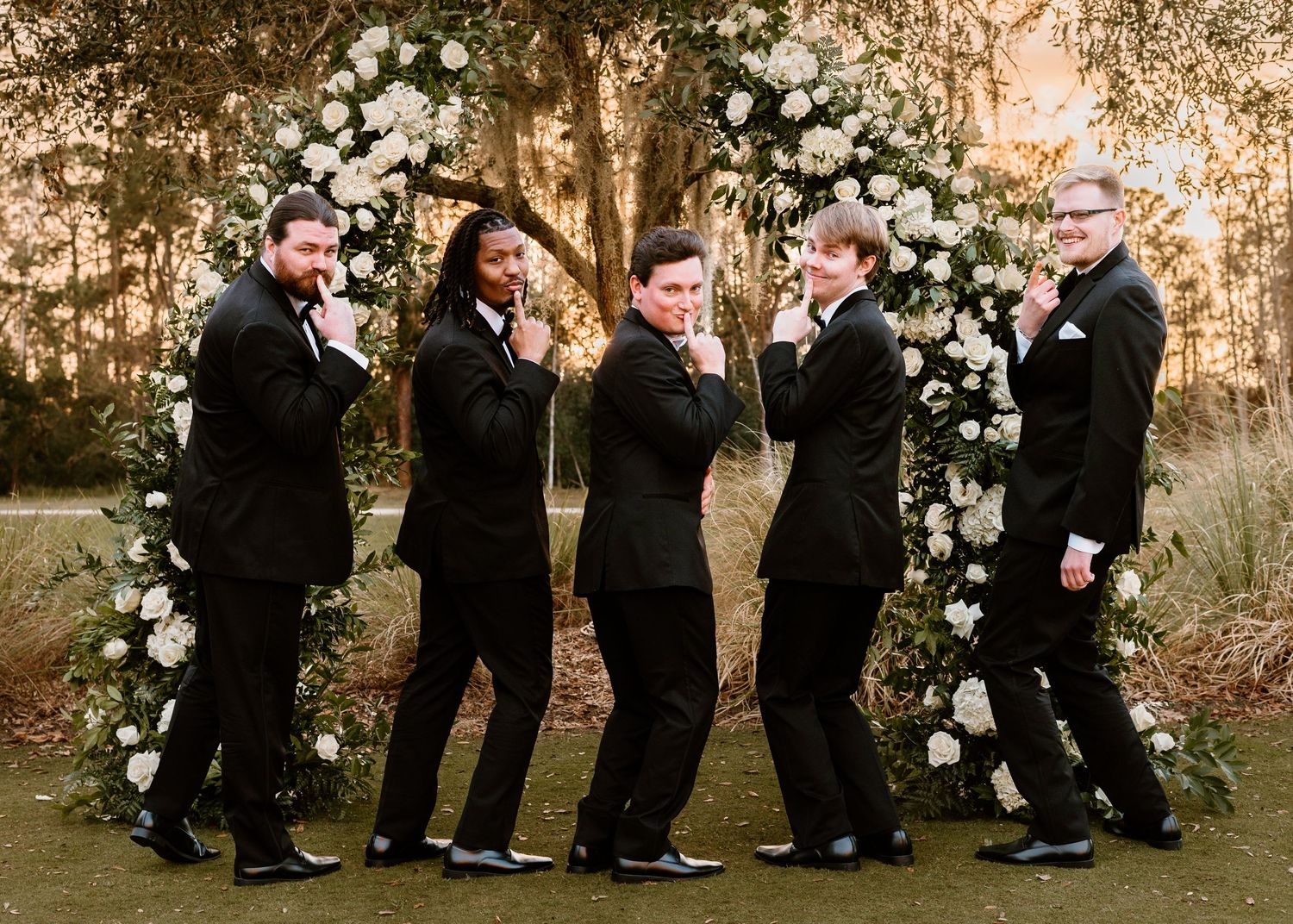 Groomsmen in black suits pose in a fun formation near a floral wedding arch.