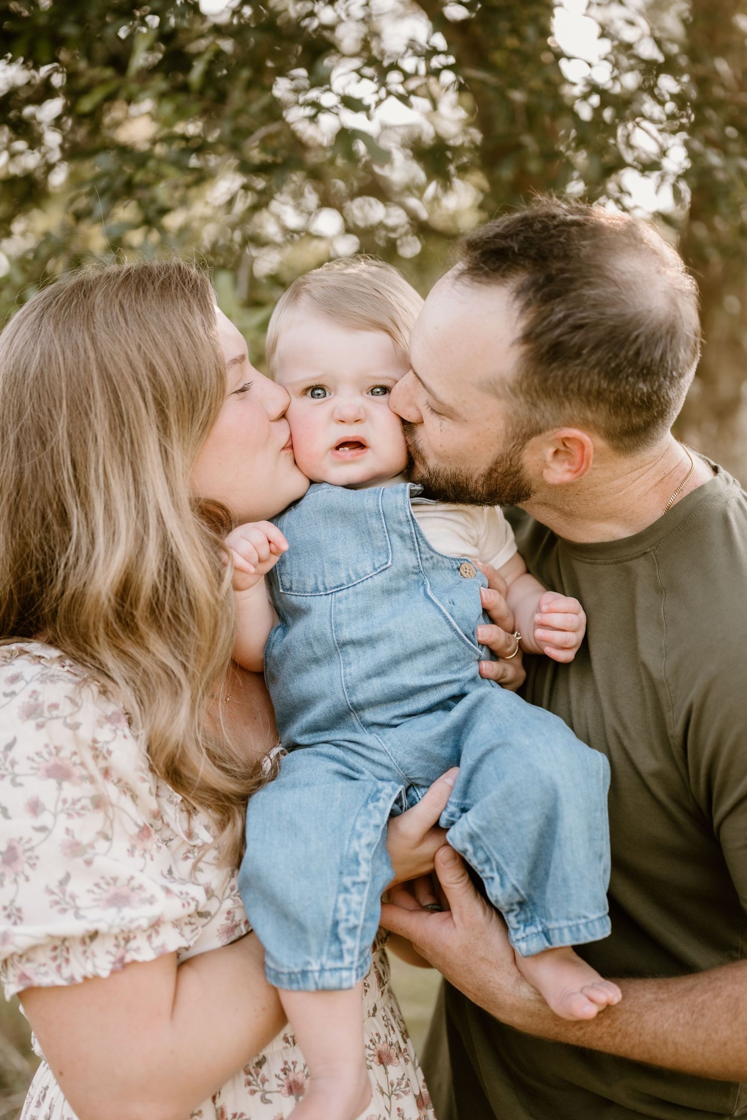 Baby in denim outfit receiving kisses from parents in outdoor setting.