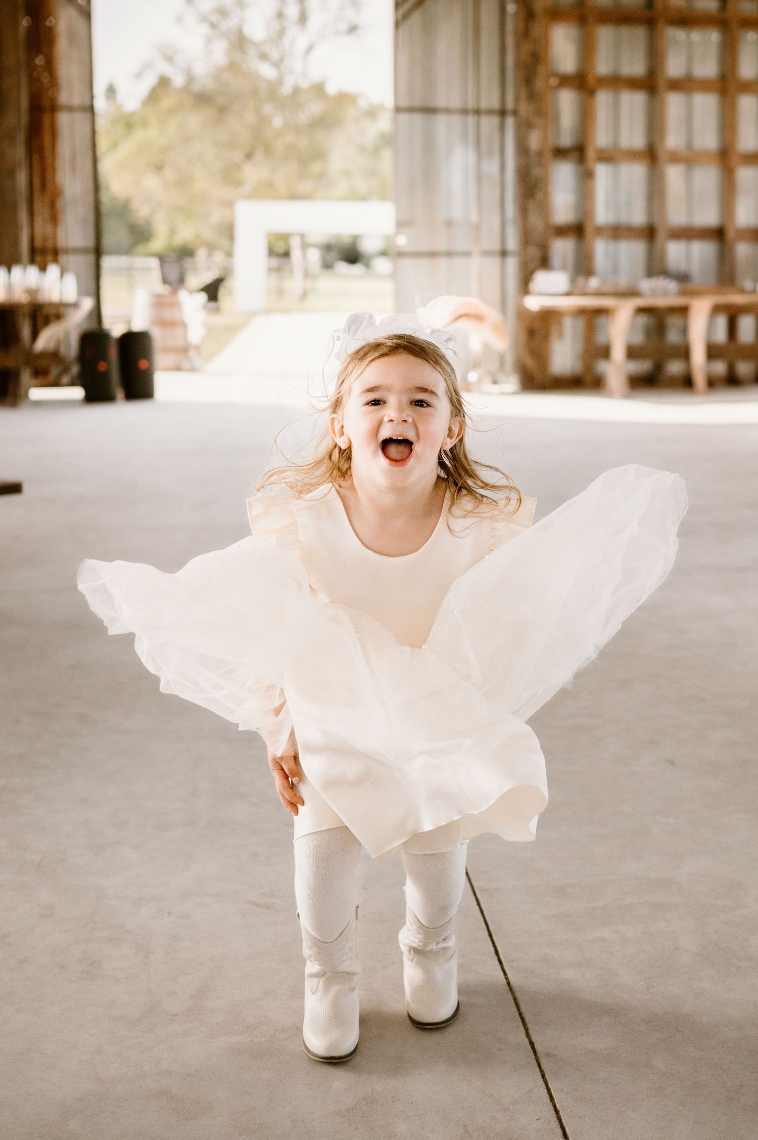 Young flower girl in white dress twirling joyfully on concrete floor.