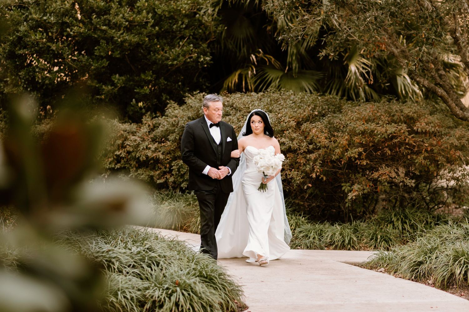 A wedding couple walks along a garden path surrounded by lush tropical landscaping.