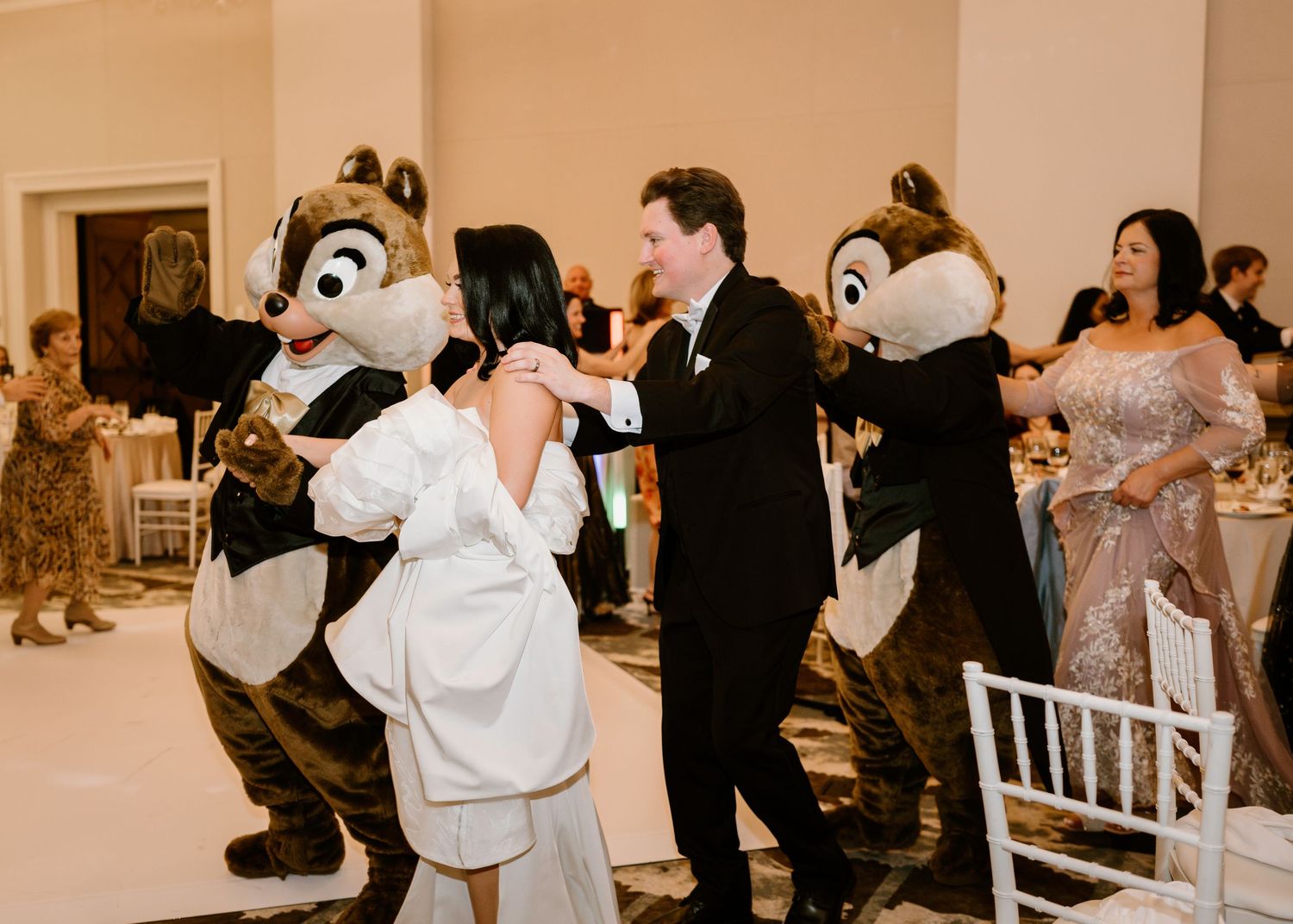 Wedding reception guests dance with Disney character mascots in a banquet hall.