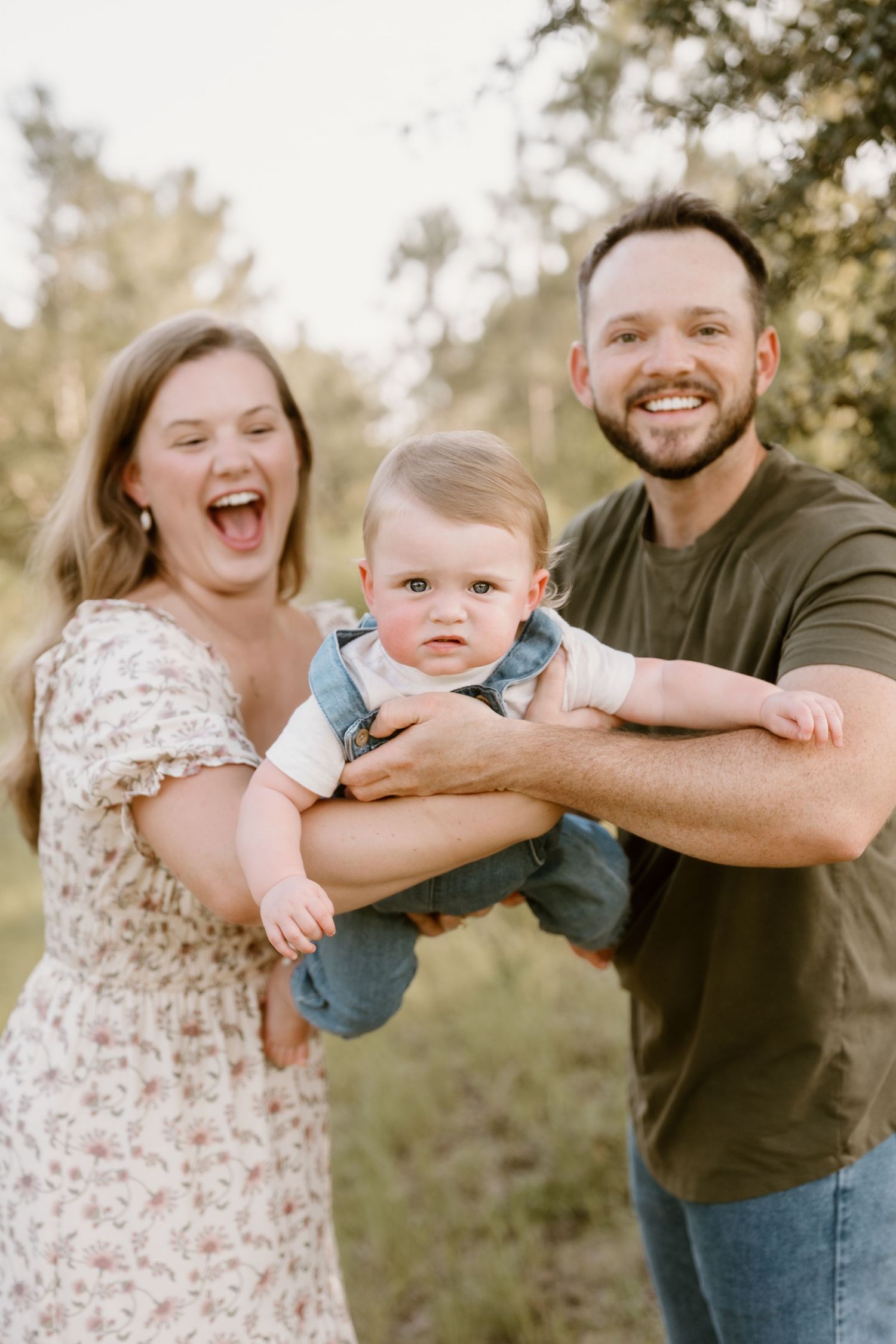 A joyful family portrait outdoors with a baby wearing denim overalls in a sunlit natural setting.