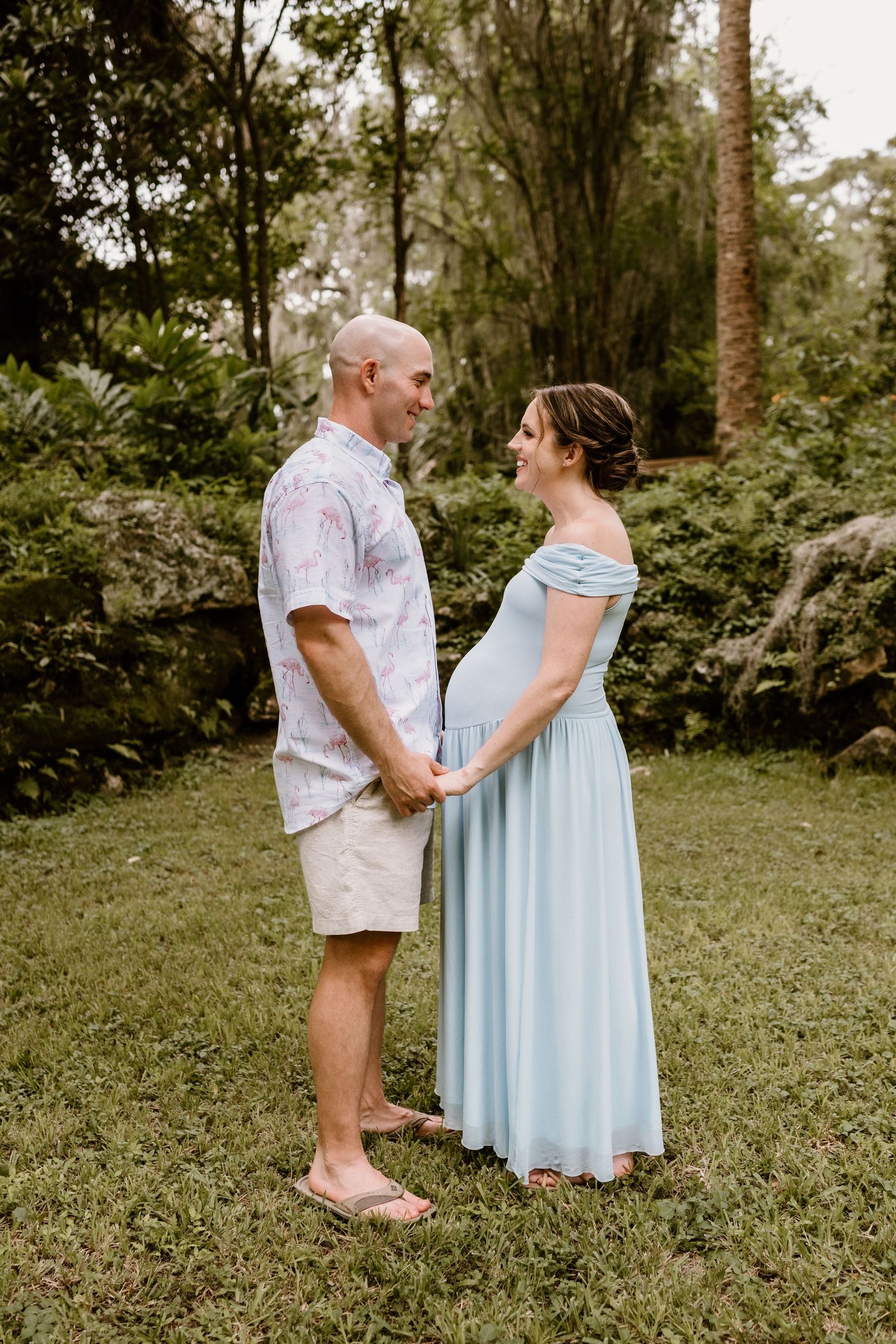 Maternity portrait of couple in coordinated outfits standing together in a green garden.