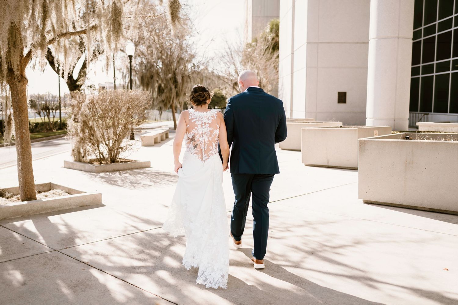 Wedding couple walking along a sunlit path with trees and building shadows.