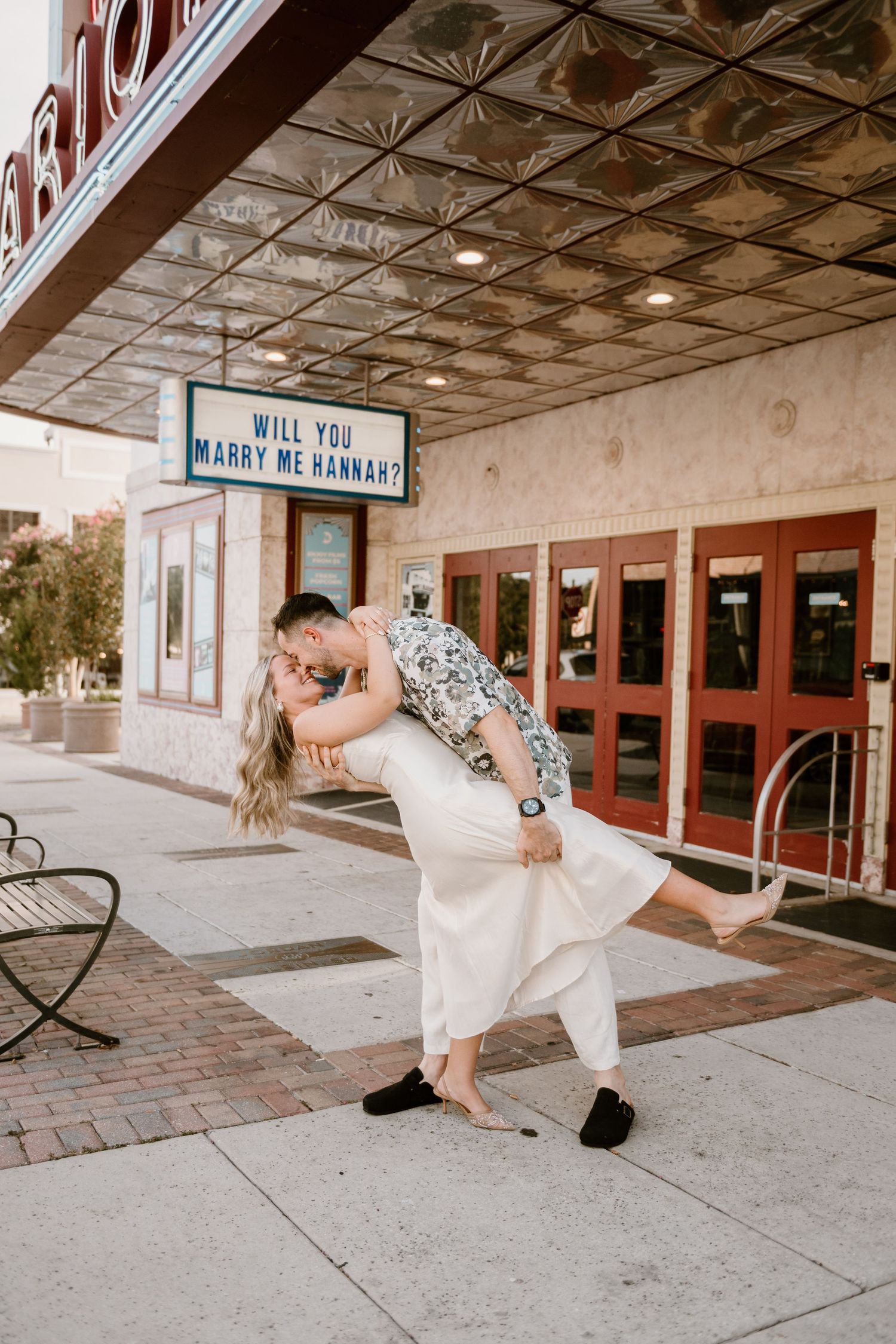A couple celebrates their engagement with dramatic poses outside a classic theatre.