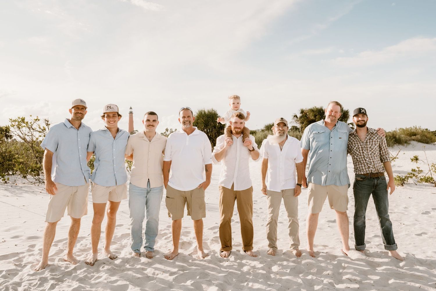 A large group poses together on a beach wearing coordinated light-colored casual outfits.