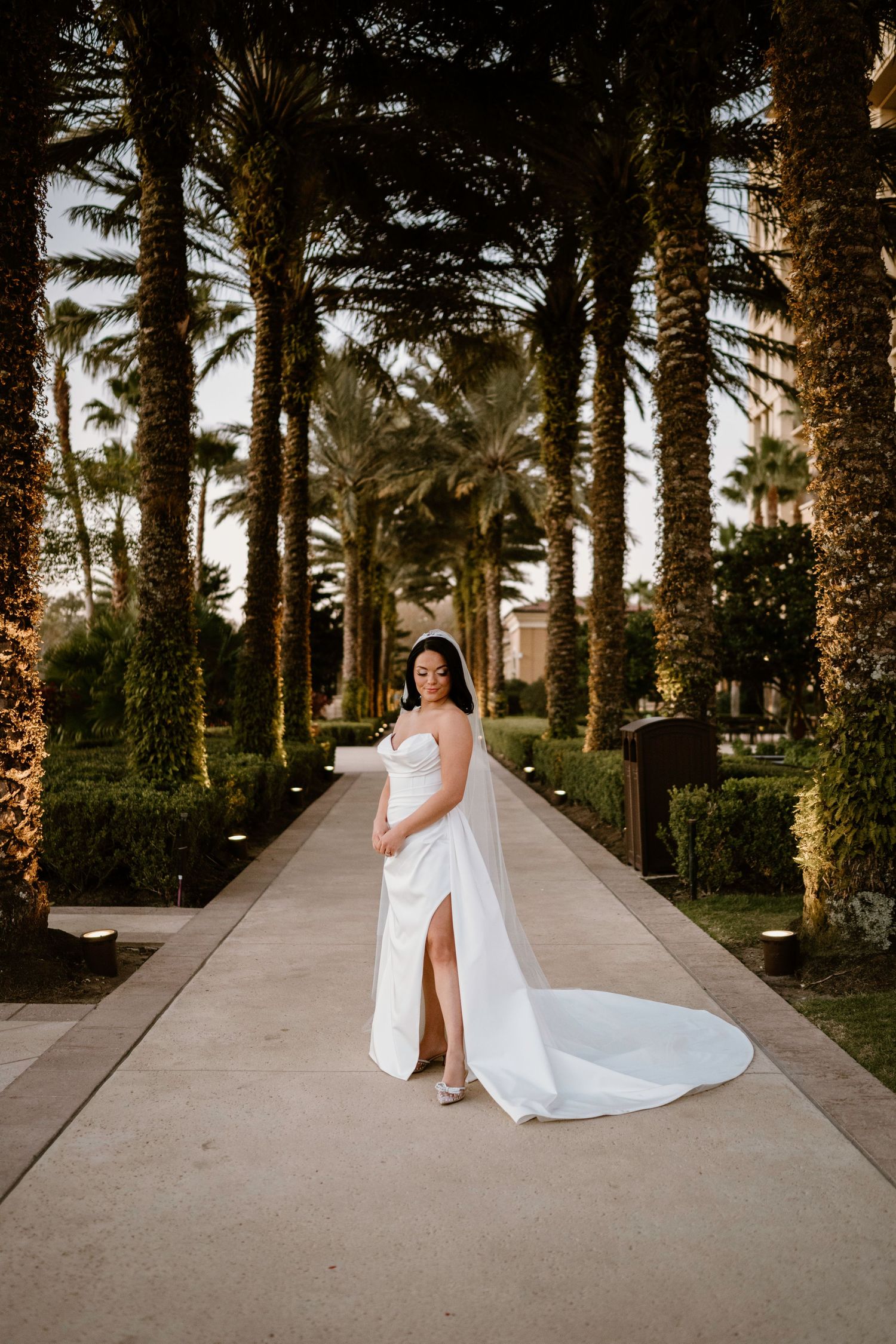 Bride in flowing white dress poses along palm-lined pathway at sunset.