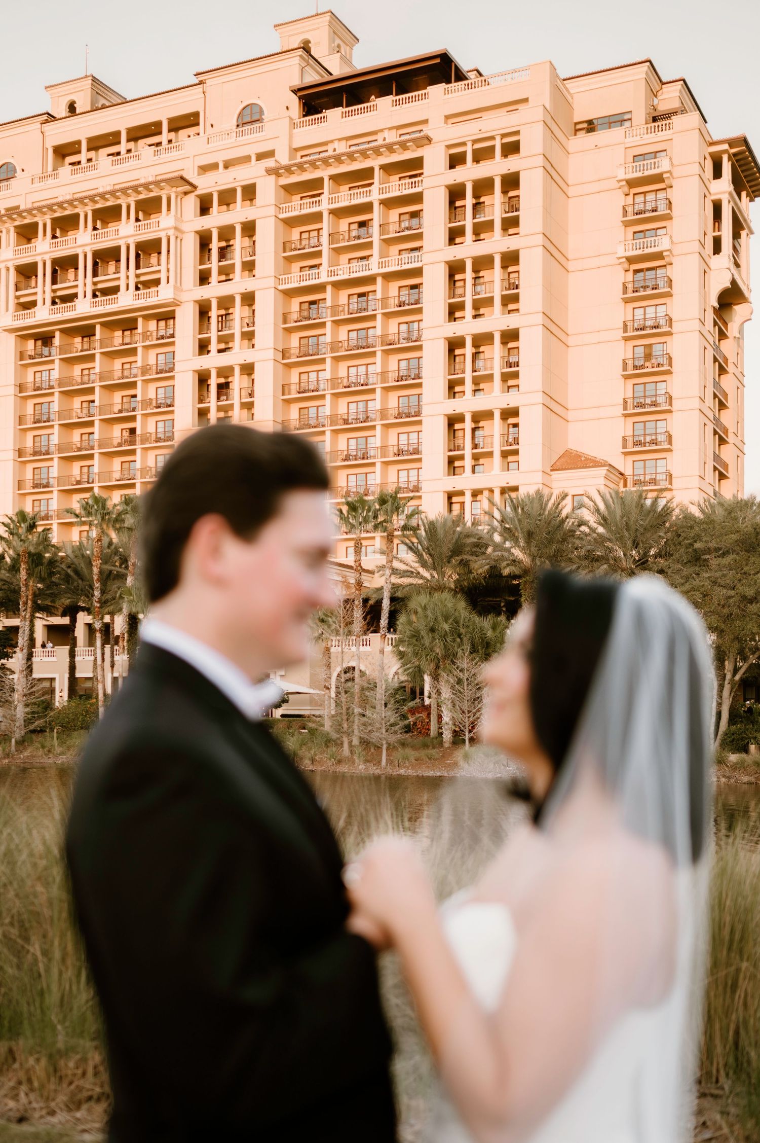 Wedding couple poses in front of elegant beachfront resort at golden hour.