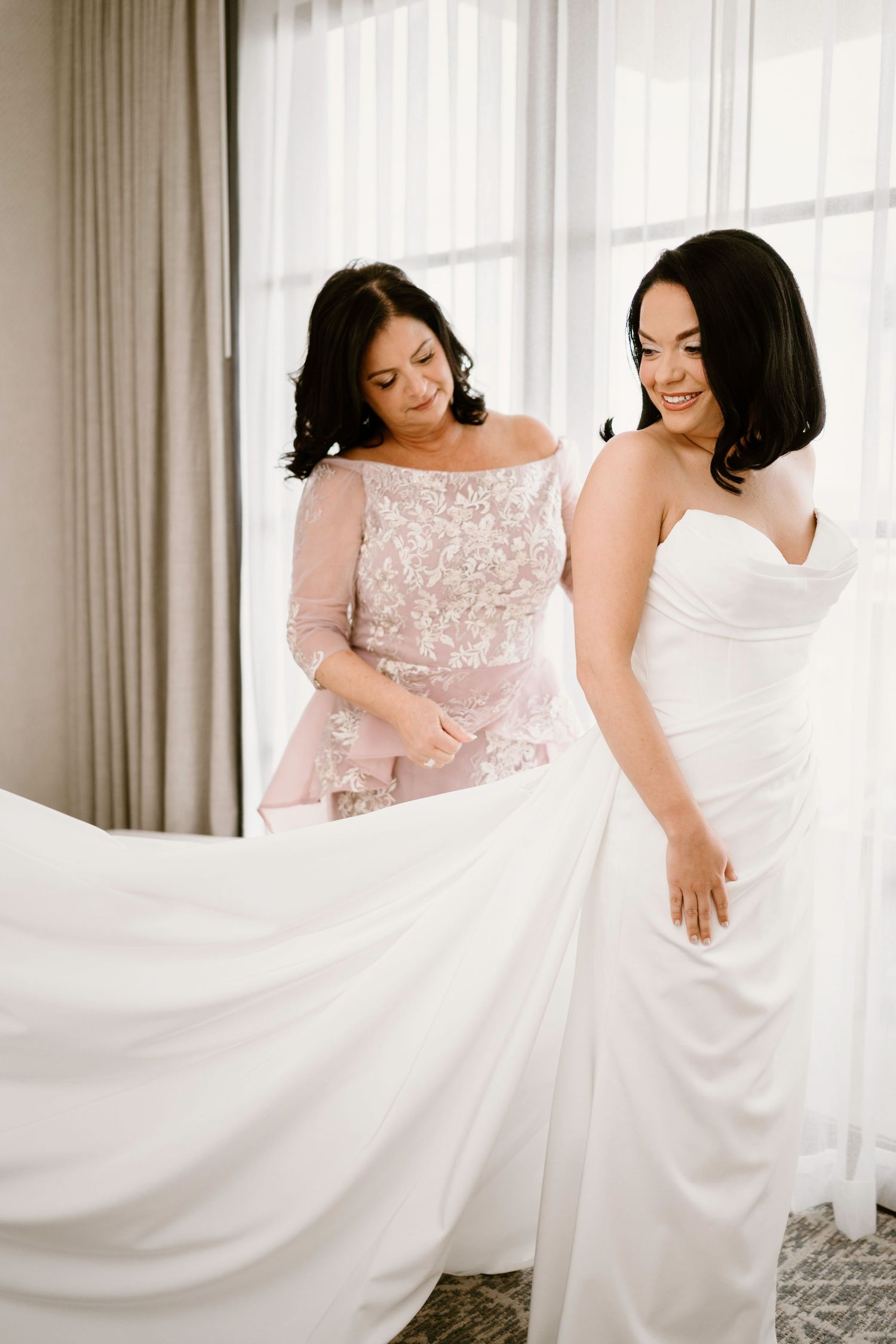 Two women in formal dresses twirl and admire flowing white fabric in a sunlit room.
