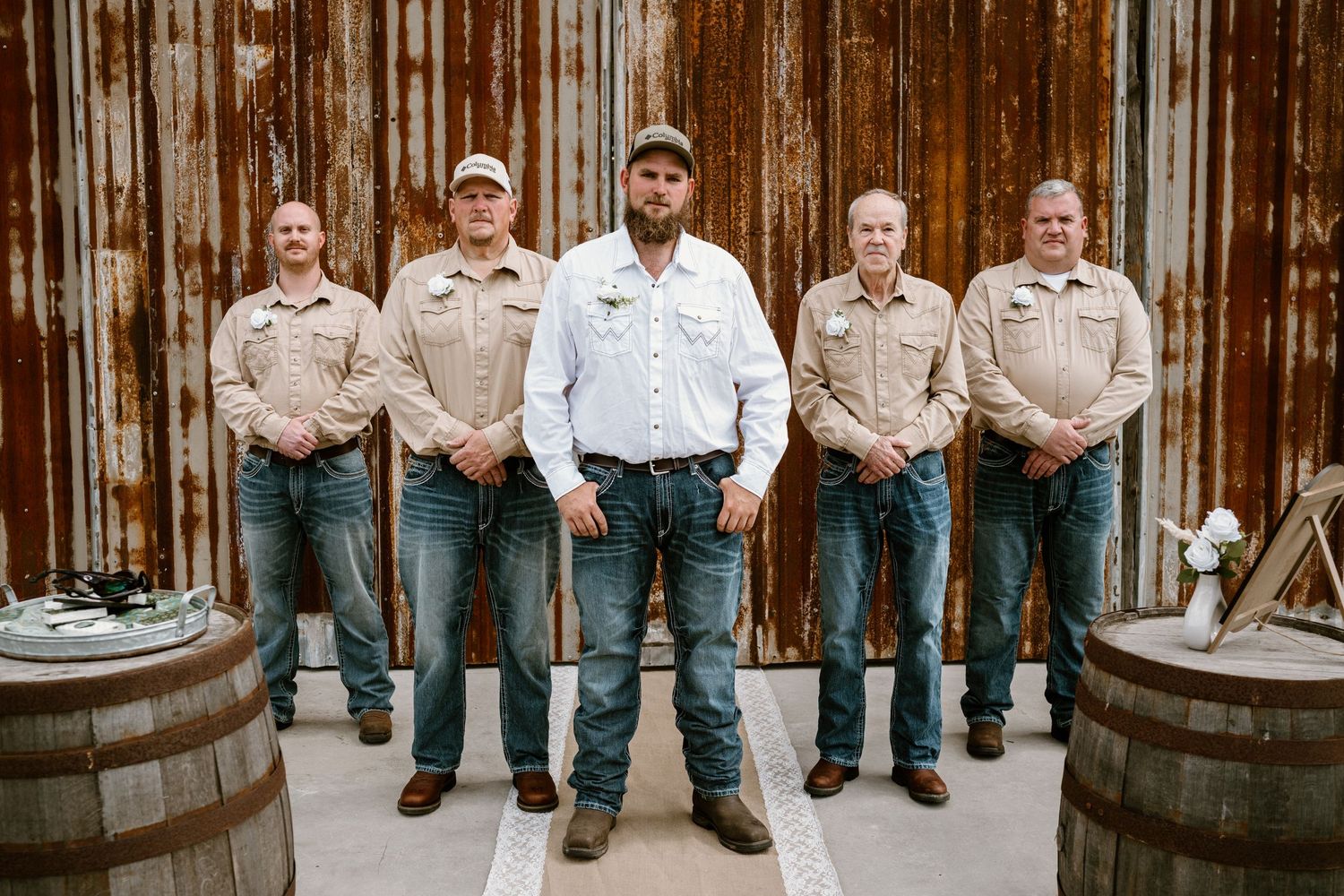 Wedding party members sitting on wooden barrels against rustic barn wall.