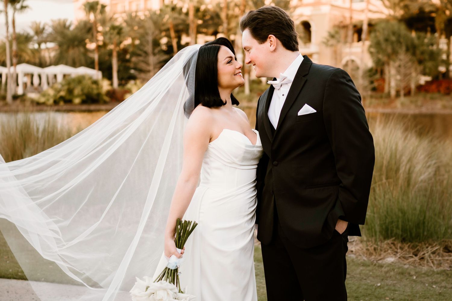 Romantic wedding couple poses at sunset with palm trees and flowing veil creating dreamy silhouettes.