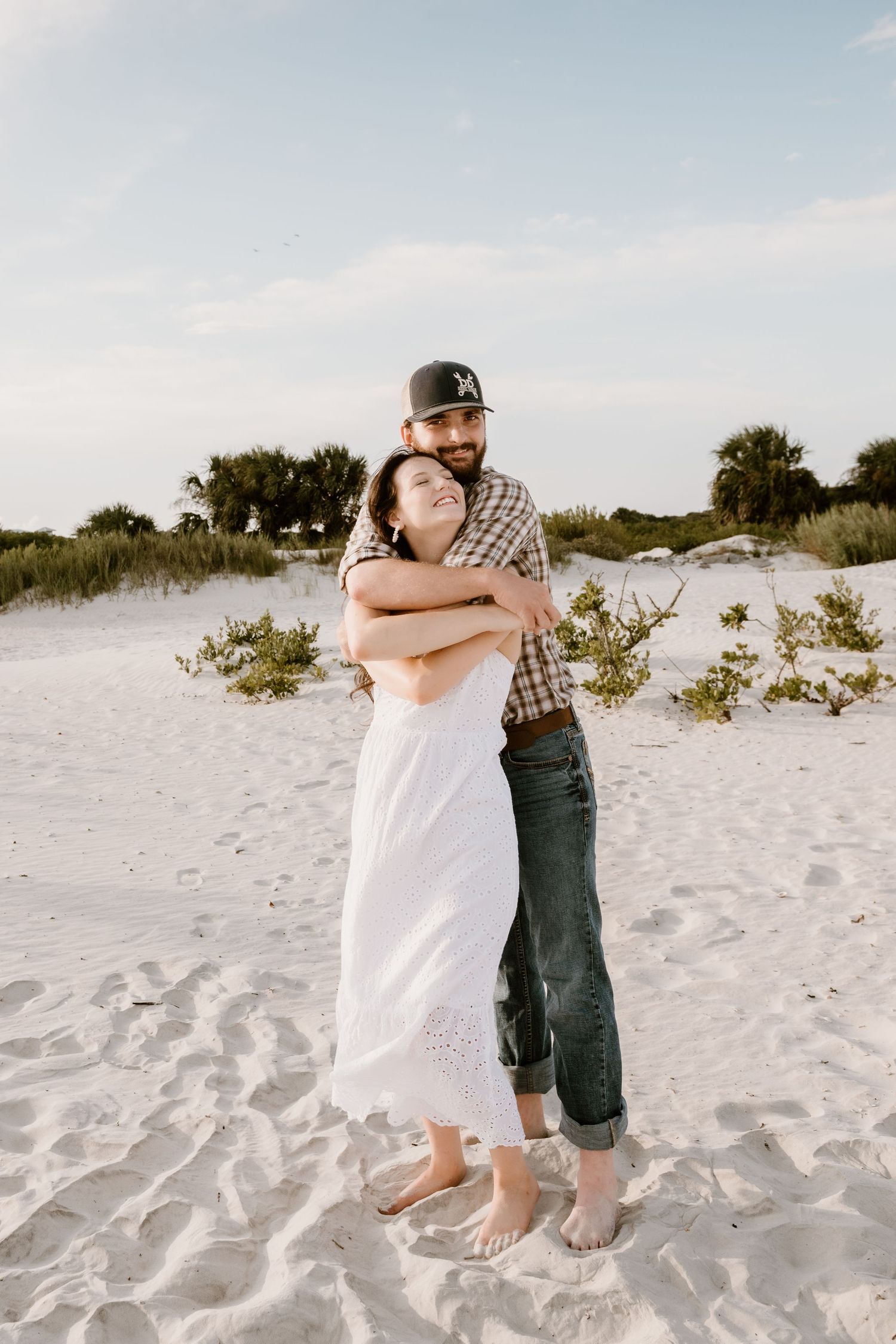 Two people embrace while dancing on a scenic beach with sand dunes and vegetation in the background.