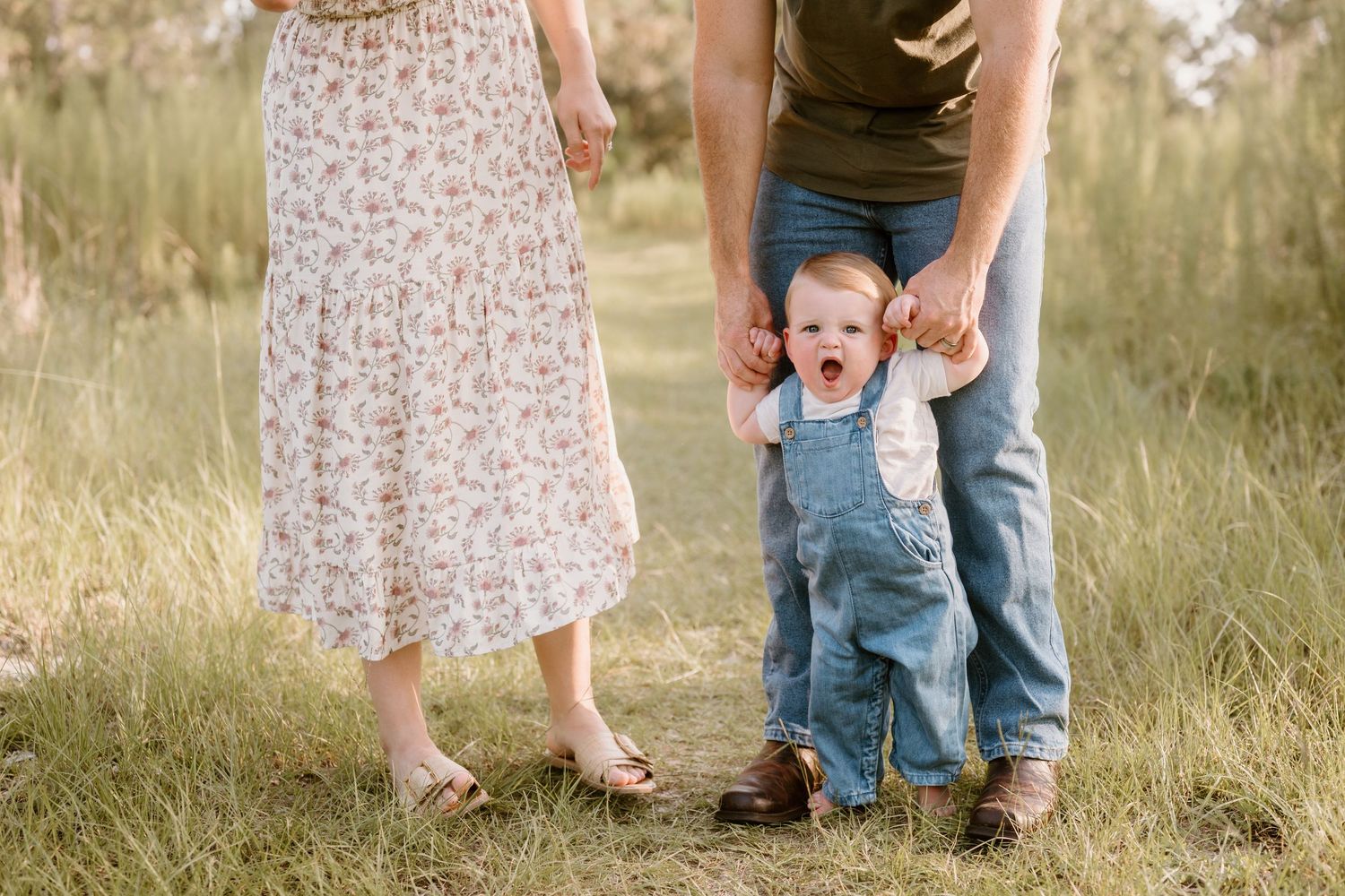 A toddler in denim overalls shows excitement while walking with parents in a grassy field.