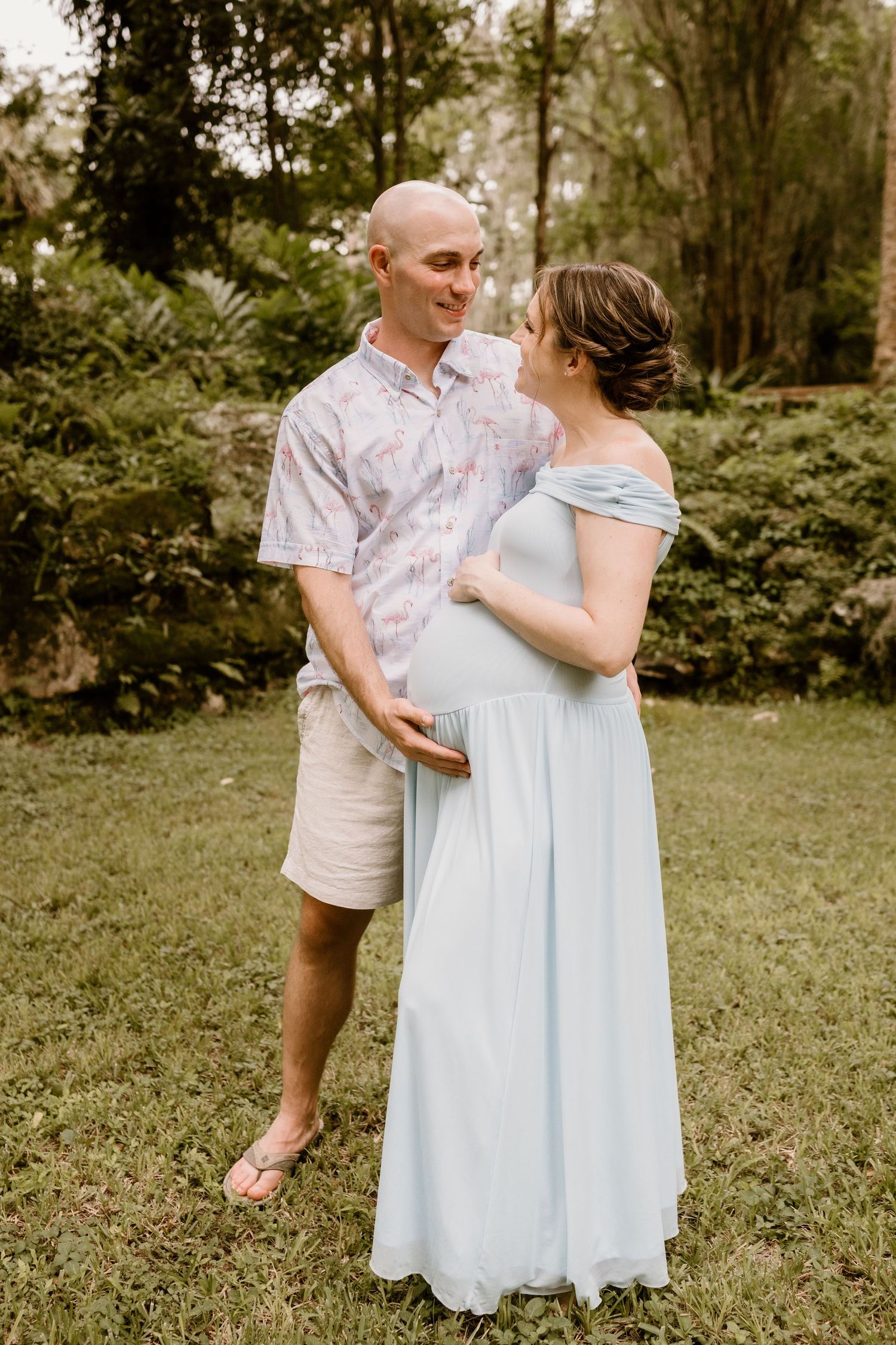 Expectant couple in light summer clothing sharing a loving moment in a lush garden setting.