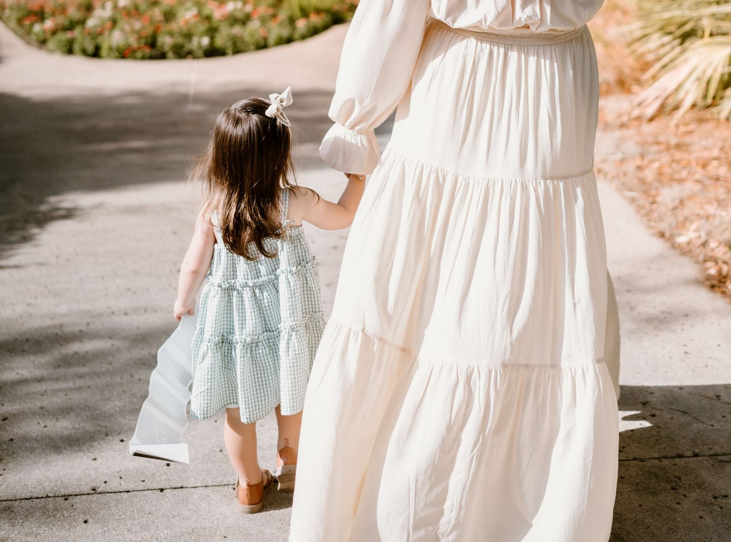 A child in a light blue dress walks alongside someone wearing a flowing white dress on a path.