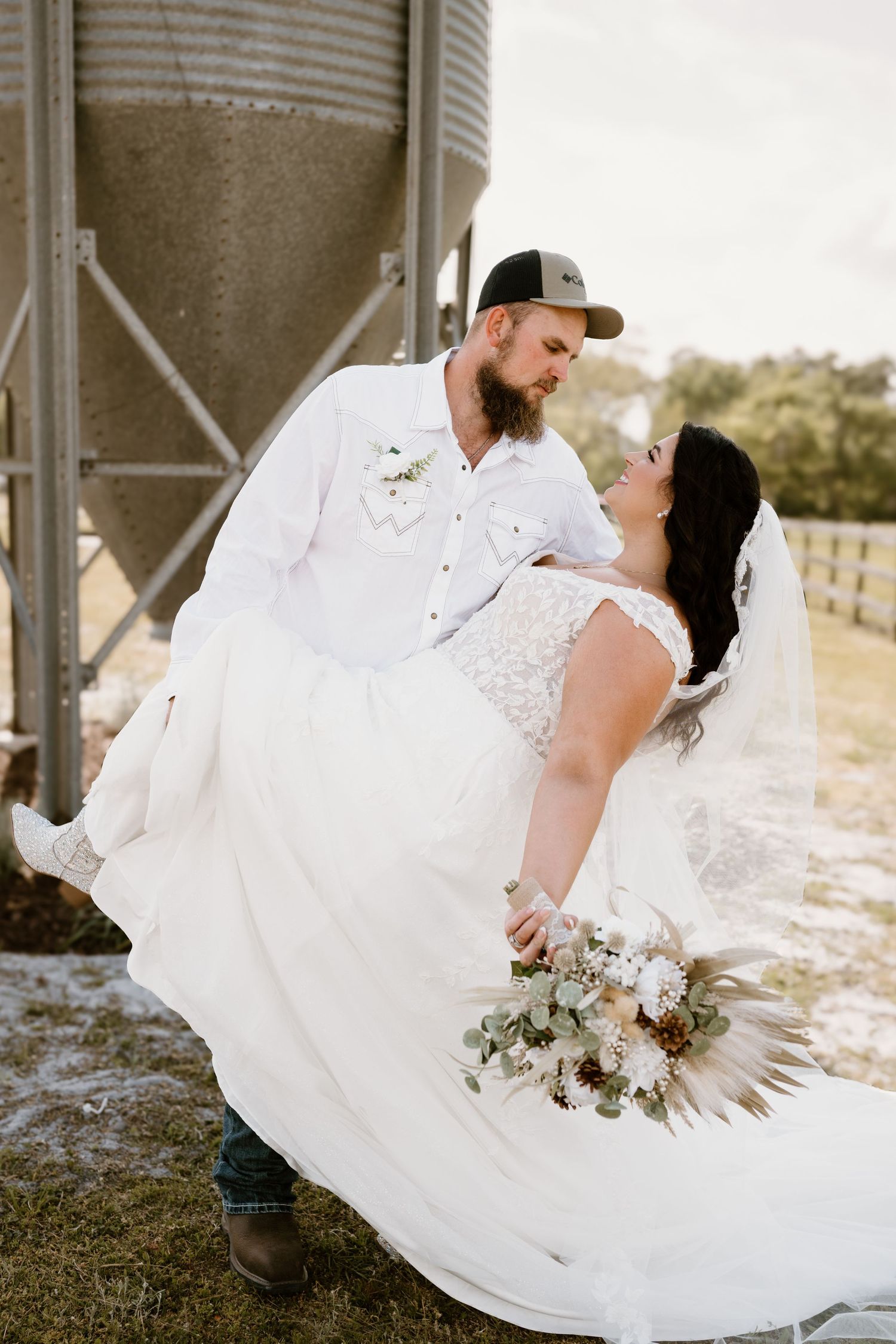 Romantic wedding portrait of couple by rustic grain silo with flowing white dress and bouquet.
