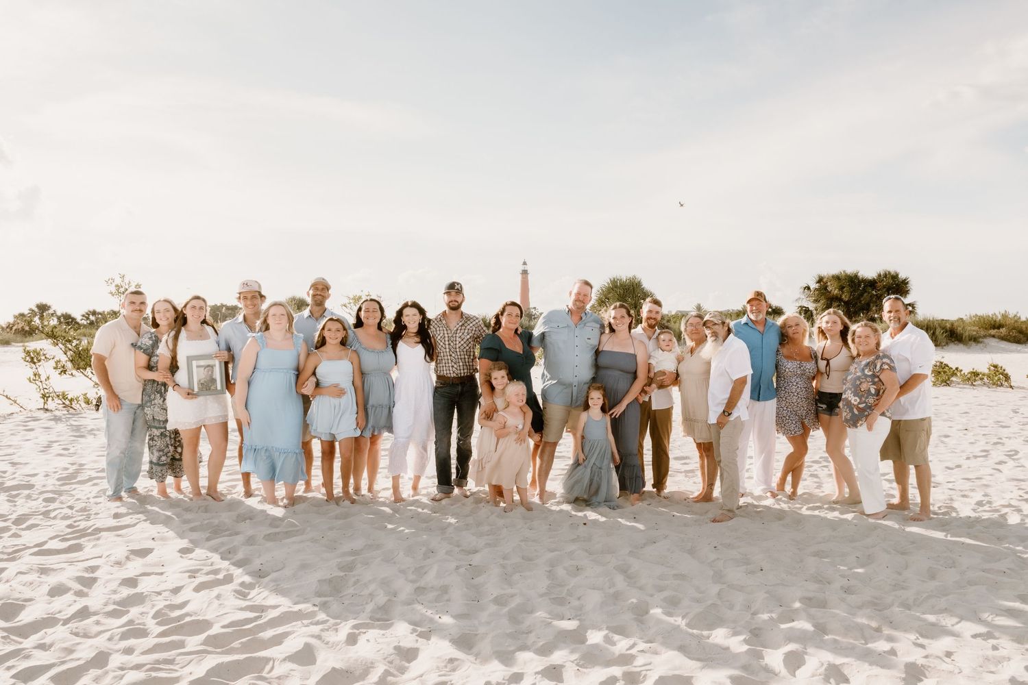 Large wedding party poses together on white sandy beach at sunset.