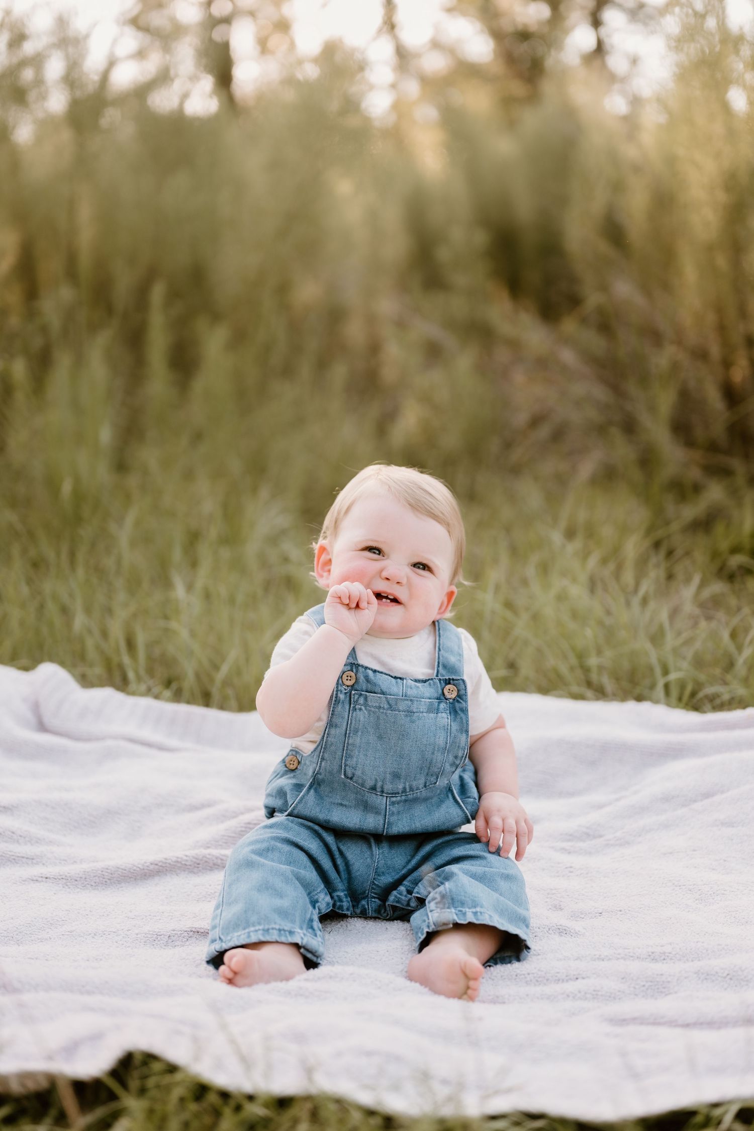 Baby in denim overalls sitting on white blanket outdoors during golden hour photoshoot.