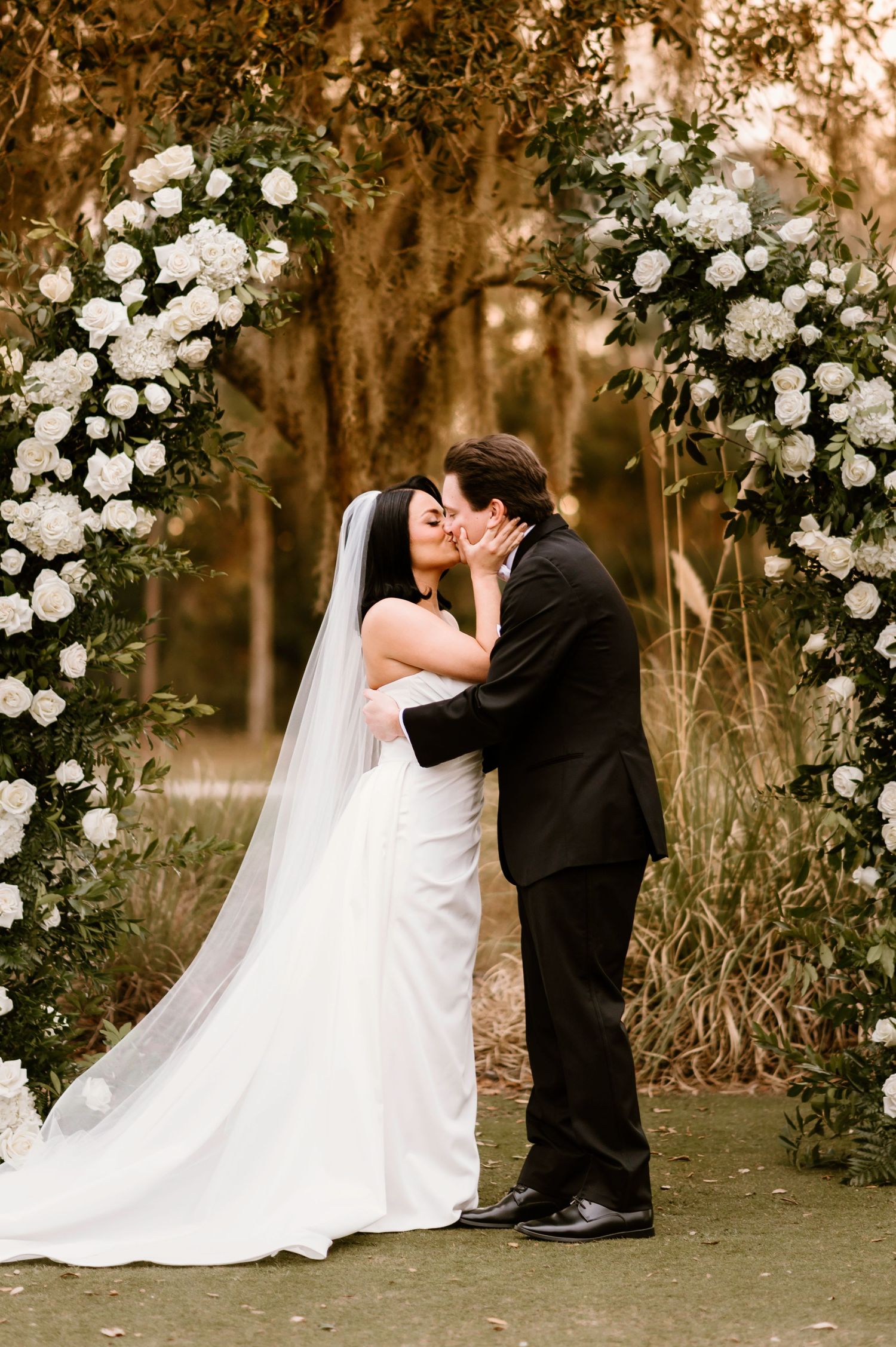 A romantic wedding moment under a floral arch decorated with white roses in a garden setting.