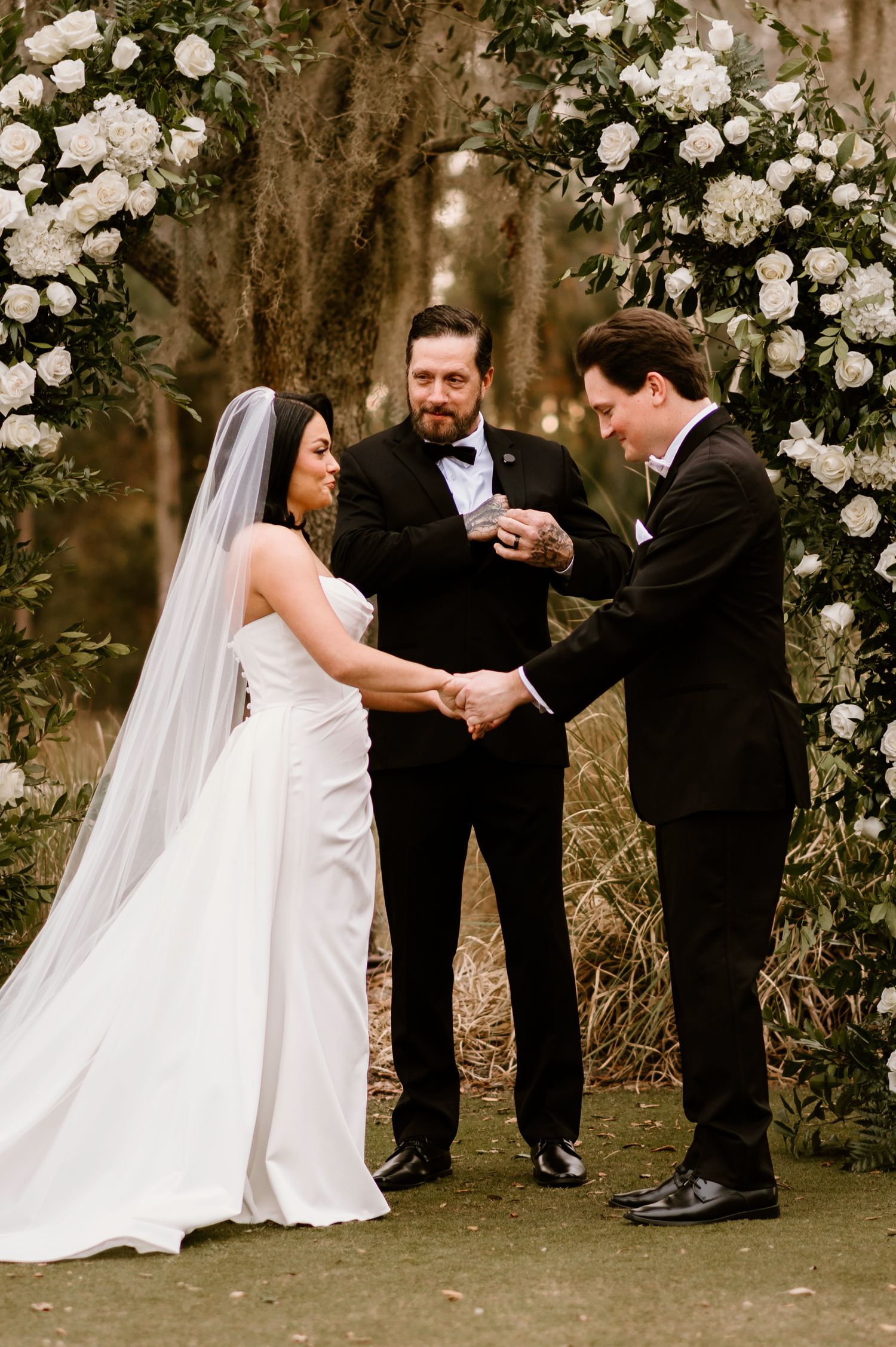 Wedding ceremony exchange of rings under an arch decorated with white roses and greenery.