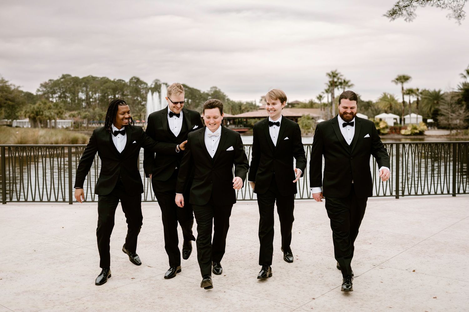 Groomsmen walking together in black tuxedos along a waterfront path.