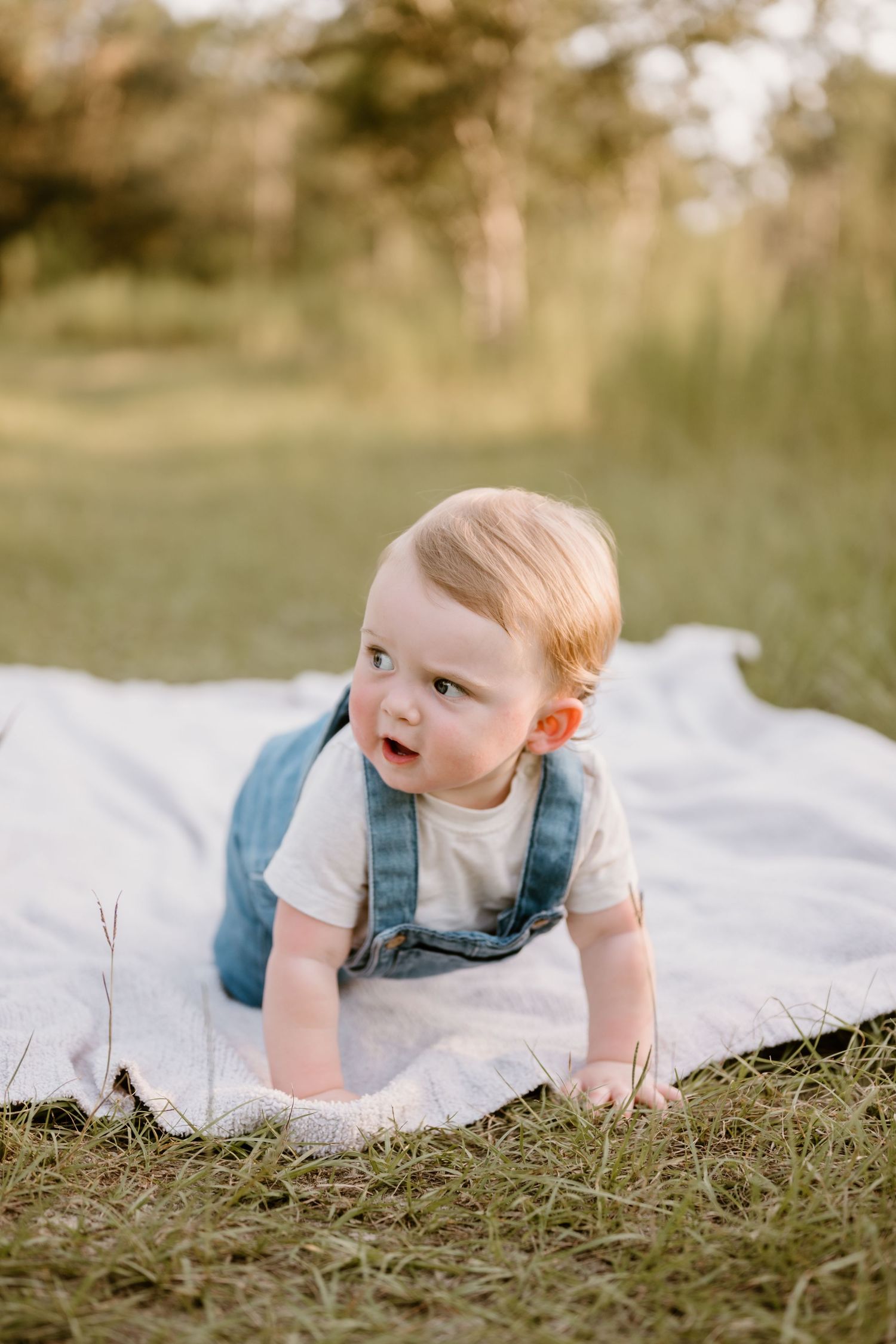 Baby in blue overalls crawling on white blanket outdoors during golden hour.