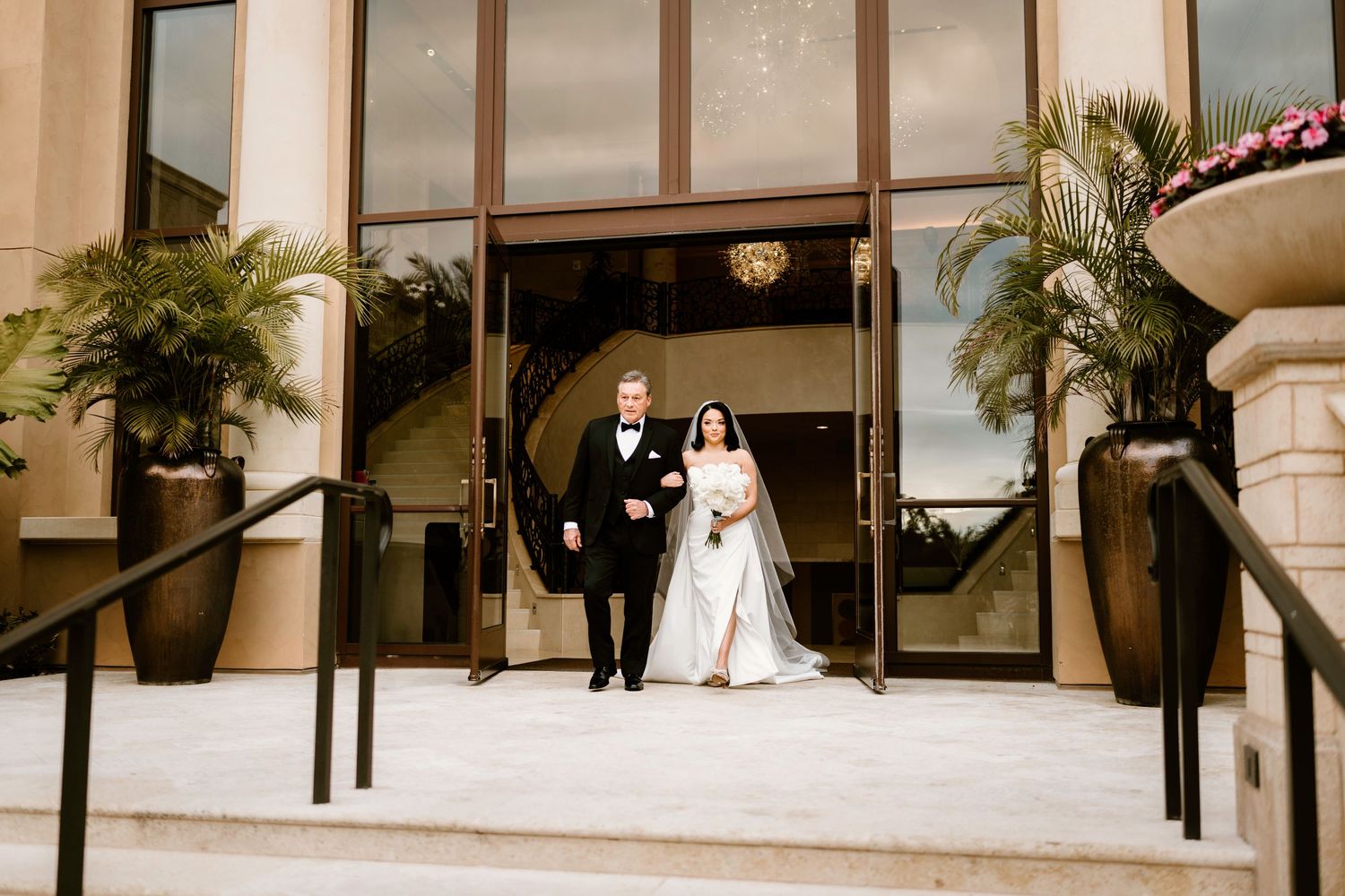 Newlyweds exit grand building entrance with stone steps and palm trees.