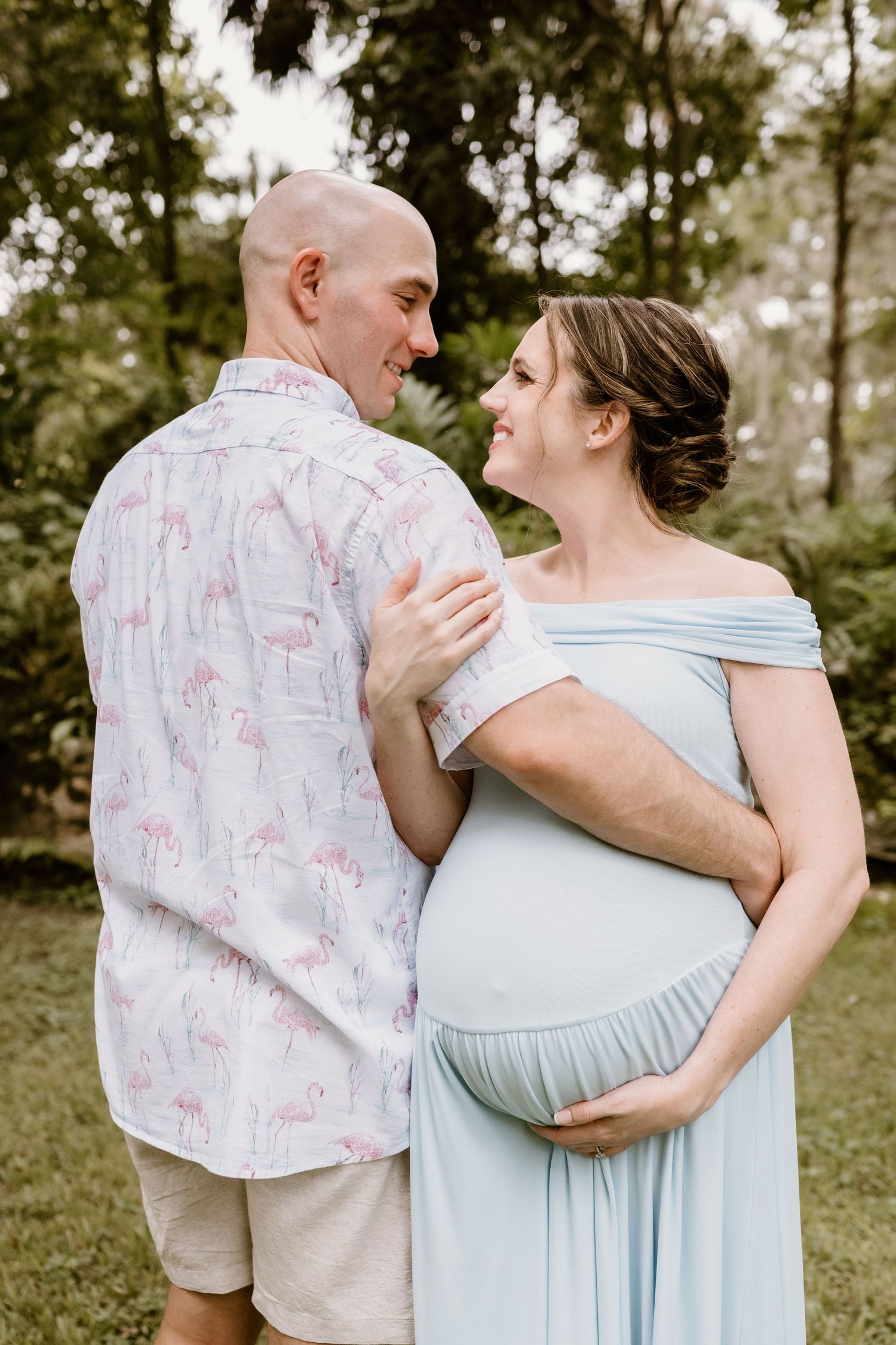 Couple in light blue maternity dress and flamingo print shirt embracing in an outdoor setting.