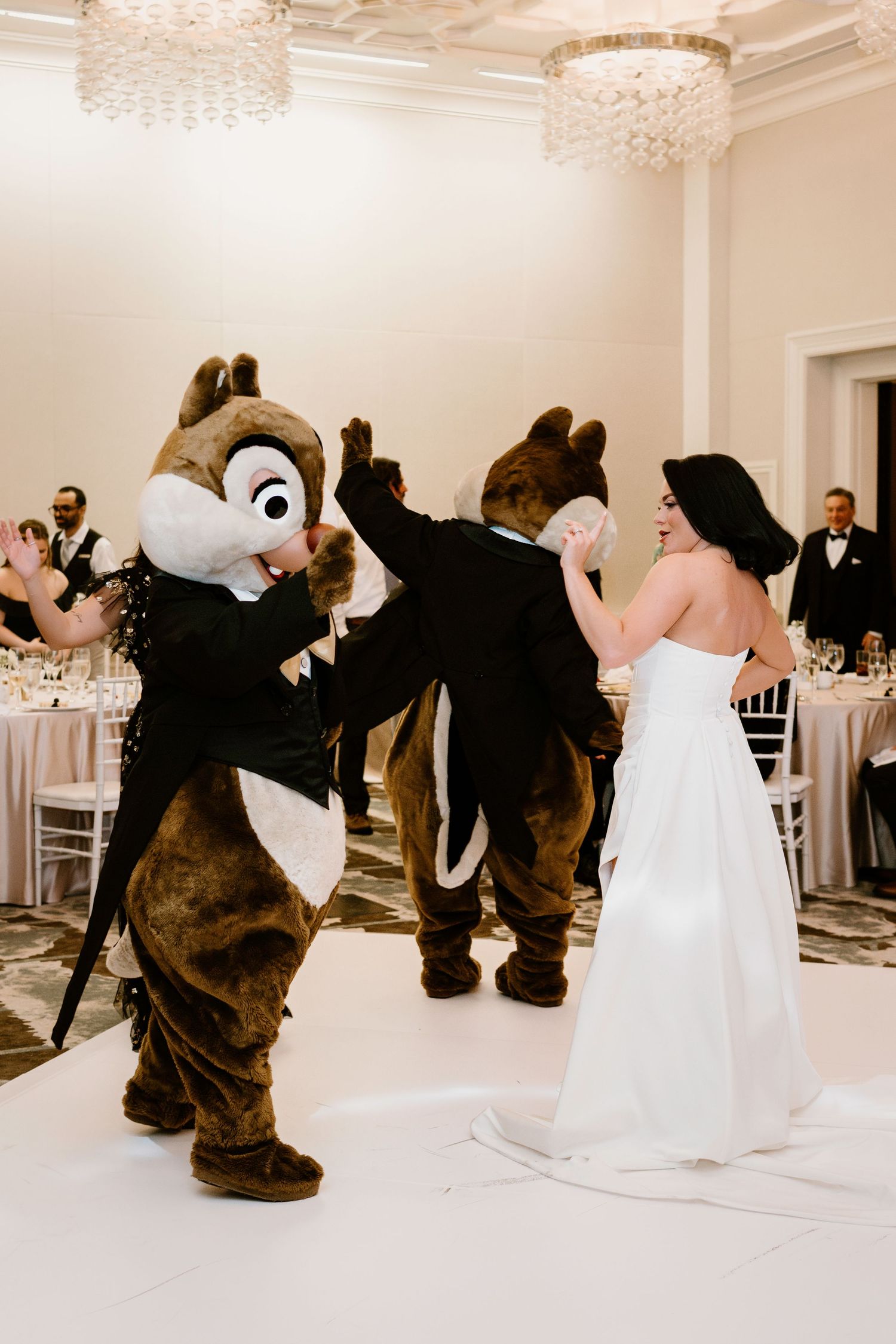 Wedding guests dance with Disney chipmunk characters under crystal chandeliers at a reception.