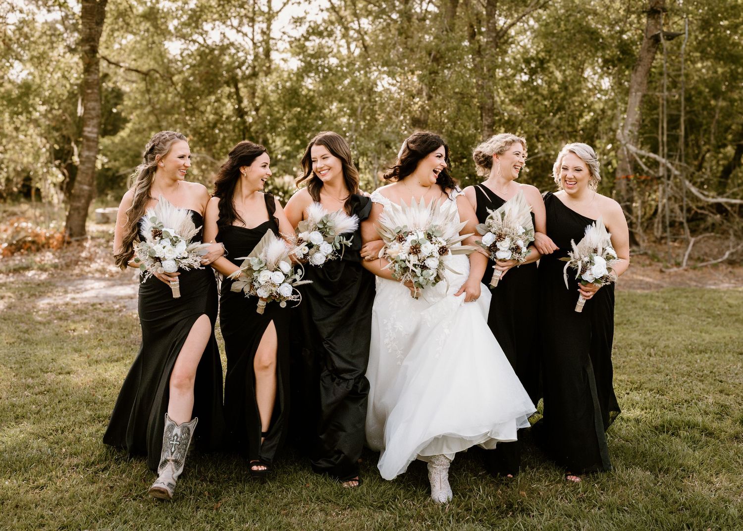 Wedding party laughing together on grass in black dresses and white gown.
