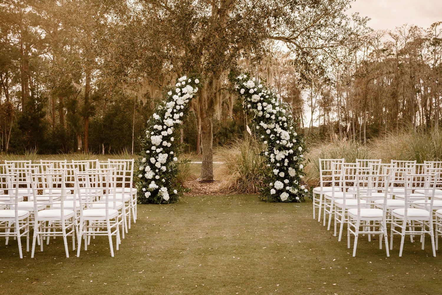 Outdoor wedding ceremony setup with white chairs and floral arch in garden setting.