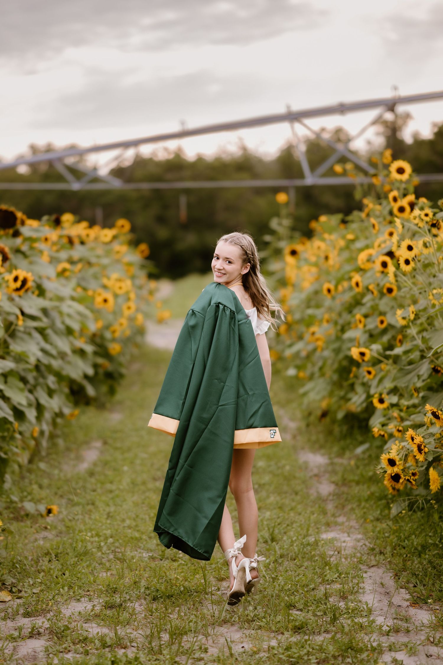 Figure carrying green coat walks along dirt path between sunflowers.